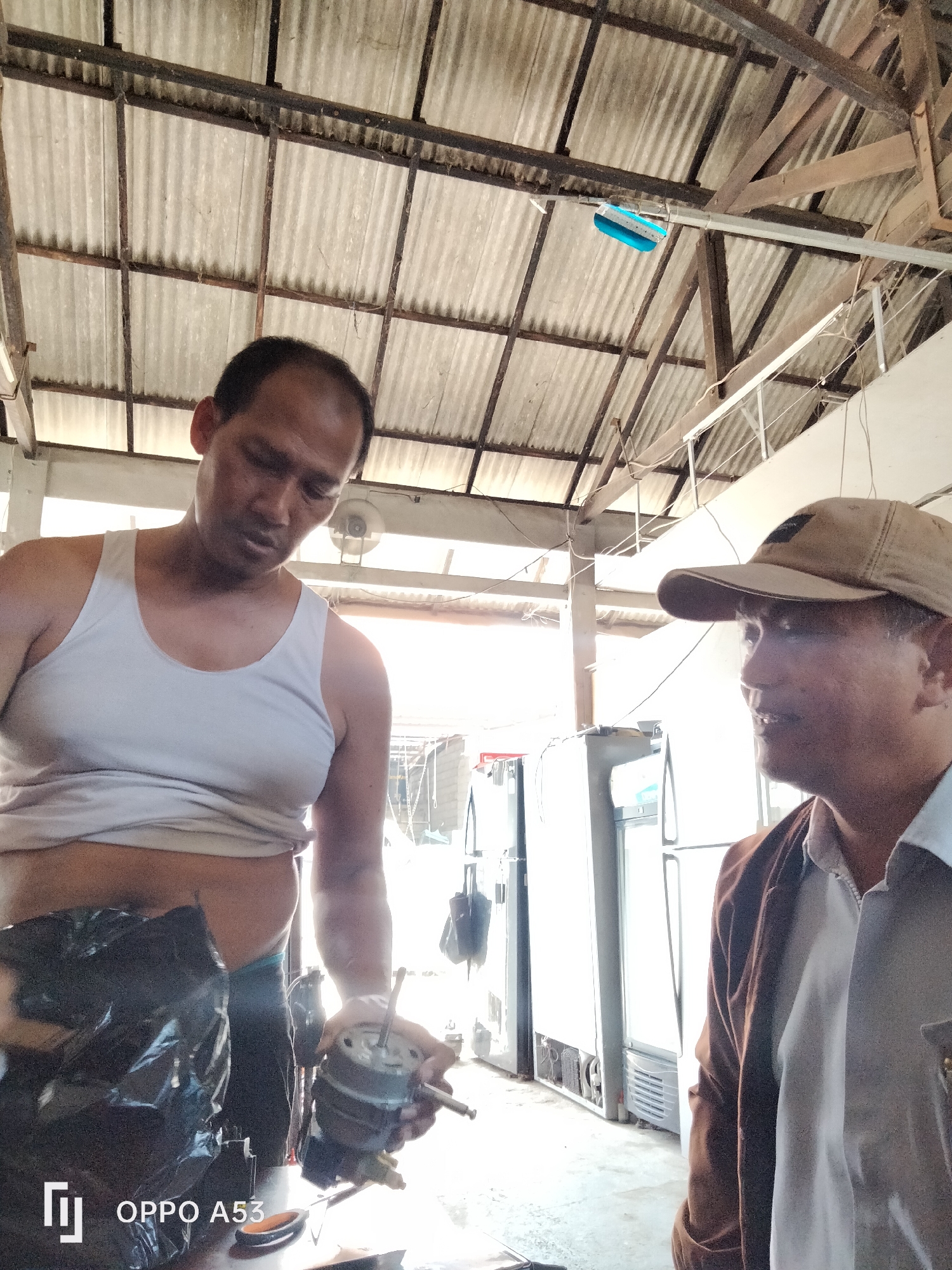 Two men are in a workshop with a corrugated metal roof supported by wooden beams. One man, wearing a white tank top, is looking down at a motor he is holding in his right hand. He appears to be in the process of repairing or examining it. Next to him, another man, wearing a brown jacket over a light-colored shirt and a tan baseball cap, is looking off to his right with a slight smile. In the background, several white refrigerators are visible, suggesting the workshop may specialize in appliance repair. The lighting suggests it is daytime, with sunlight streaming in, creating bright highlights and some shadows. The overall environment appears to be a functional, possibly outdoor or semi-outdoor, workspace.