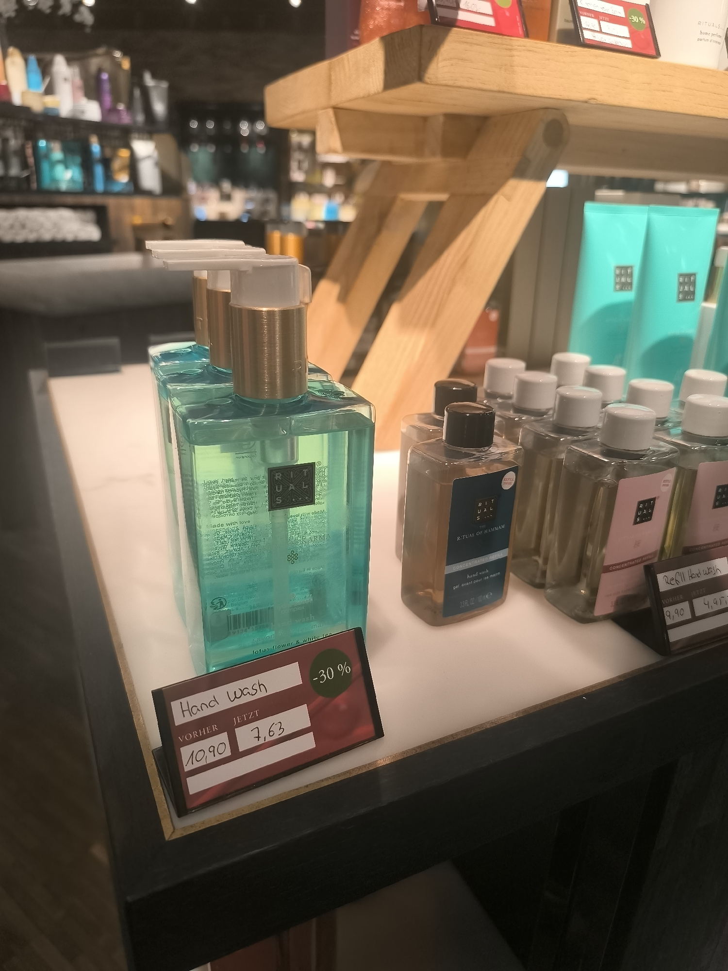 An eye-level, medium shot captures a retail display of hand wash products, illuminated by artificial lighting. The scene is set in an interior space, likely a store, with shelves of various cosmetic items visible in the blurred background. In the foreground, a dark red price tag is prominently displayed, indicating a "Hand Wash" with a previous price of "10,90" and a current price of "7,63", along with a "-30%" discount. To the right of this tag, another price tag shows "9,90" and "4,95".
The main subjects are several clear plastic bottles of hand wash. Two large, tall bottles filled with a turquoise liquid are positioned to the left. Each has a gold pump dispenser and a label with the "RITUALS" logo. To the right of these, a line of smaller, square bottles filled with a brownish liquid, also with "RITUALS" branding, are arranged. These bottles have white pump dispensers. Further to the right, more bottles with pinkish liquid are visible. The overall impression is one of a well-organized product display in a commercial setting.