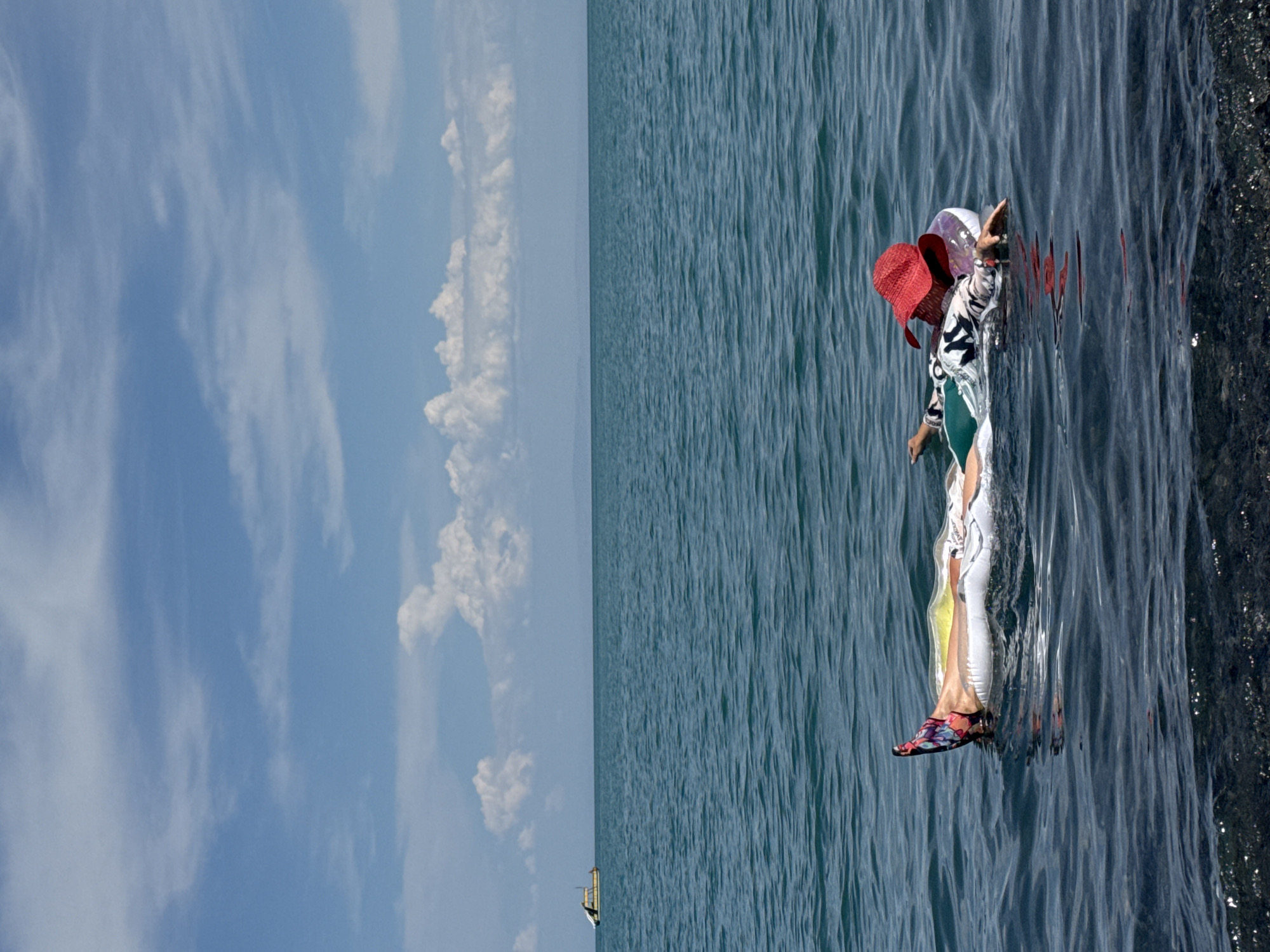 A person is relaxing on an inflatable raft in the water, wearing a red hat and a patterned shirt. The raft is floating on a body of water that appears to be a lake, with a clear blue sky and scattered clouds above. In the distance, a small boat is visible on the horizon. The foreground shows a dark, rocky shore. The overall scene suggests a peaceful summer day in a natural setting. This is likely the lake shore in Ақши, Kazakhstan, a region known for its scenic beauty and recreational opportunities.