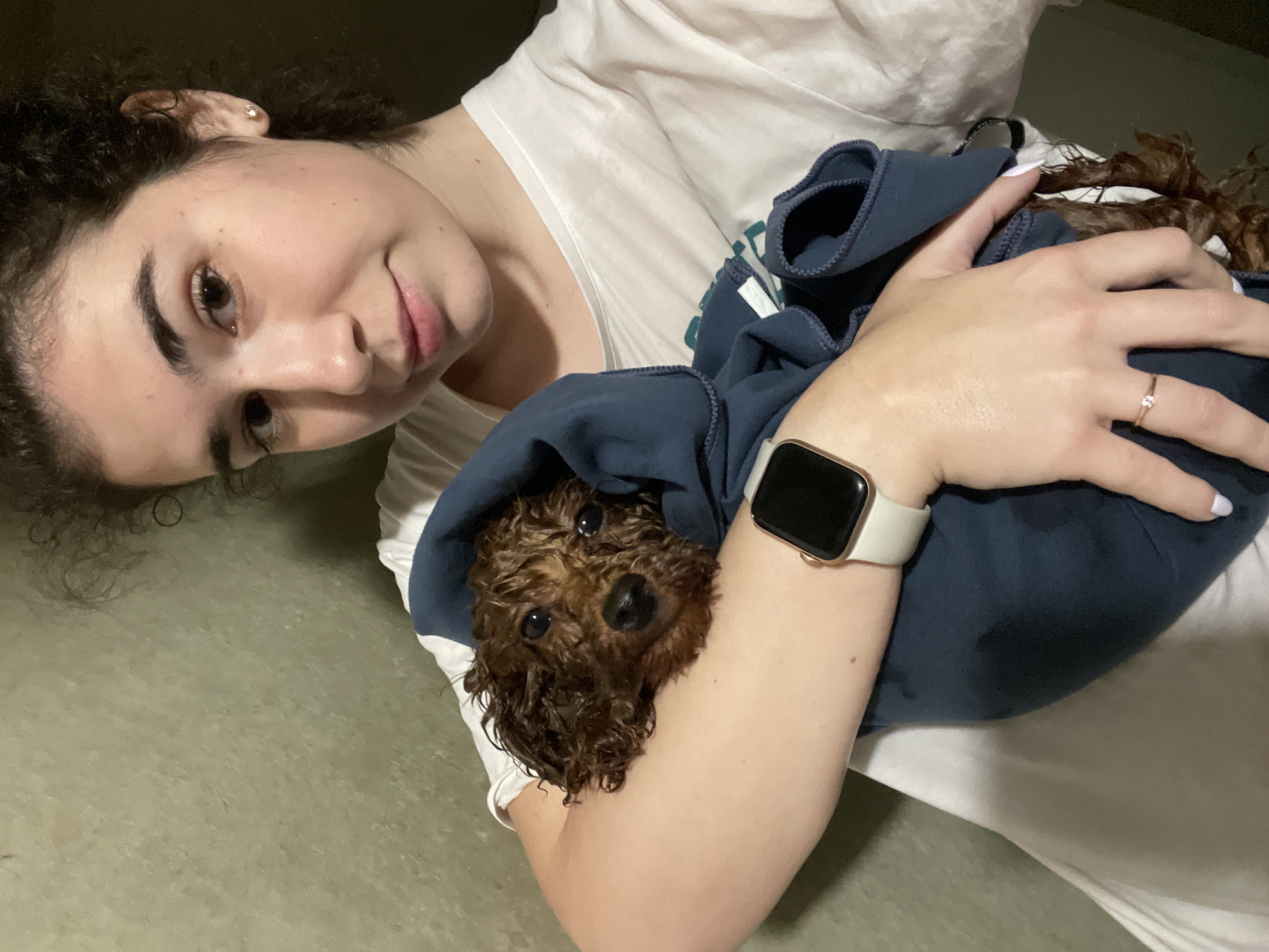 A woman with dark curly hair and fair skin is holding a small, wet, brown poodle puppy wrapped in a blue cloth against her chest. The woman is wearing a white T-shirt, a ring on her finger, and a smartwatch with a light-colored band. The puppy is looking directly at the camera. The background is a plain, light-colored floor. The image was likely taken indoors and is not identifiable as being in Sofia, Bulgaria.