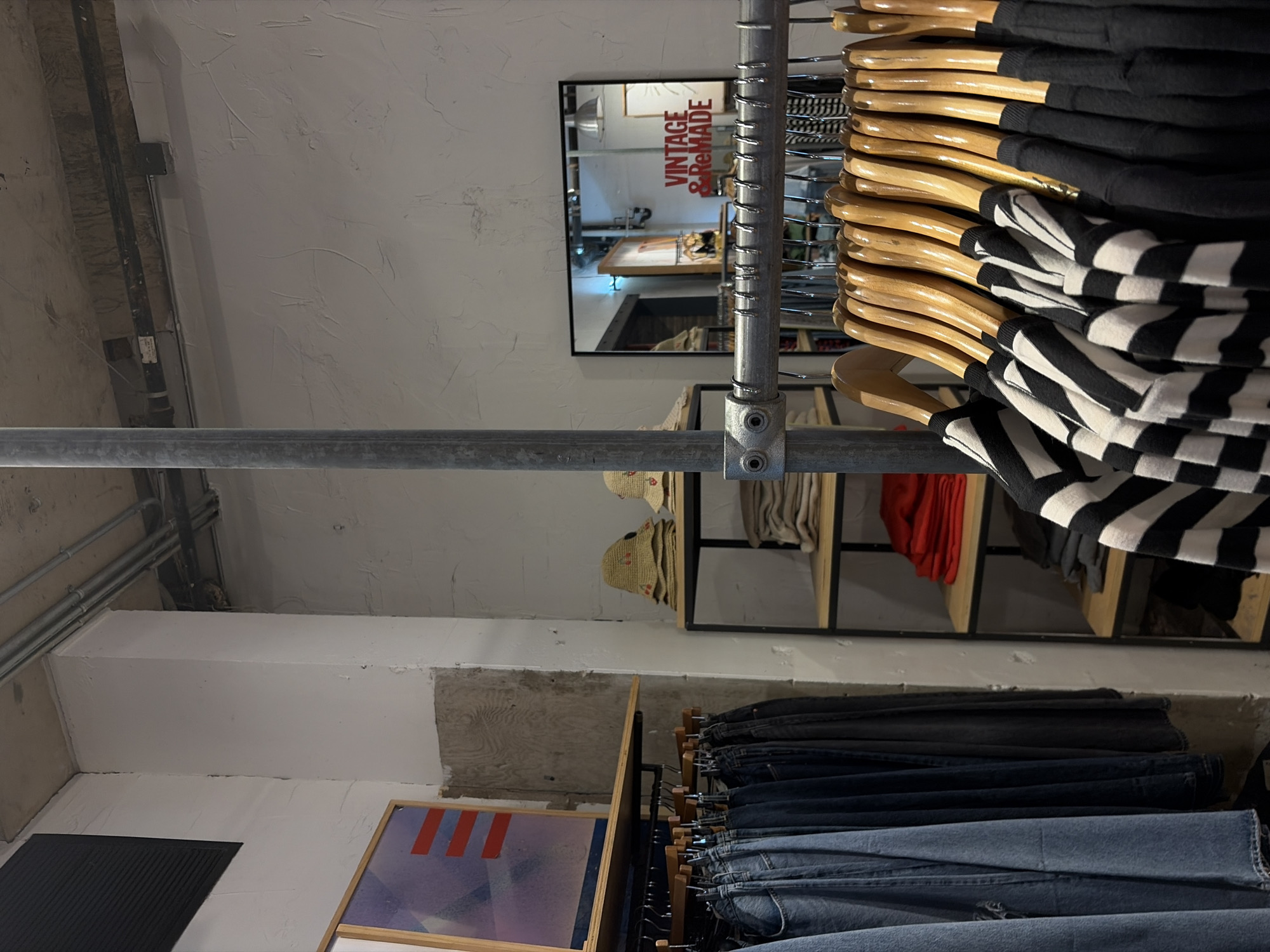 The image shows the interior of a retail clothing store, possibly located in Austin, Texas, based on the presence of "Vintage & ReMade" signage. Clothes are hanging on racks, with hangers visible. A mirror is mounted on the wall reflecting the interior of the store. Shelves hold folded items, including clothing and hats. A framed graphic or artwork leans against the wall. The environment is brightly lit with plain ceiling fixtures. Visible are metal pipes and ductwork along the ceiling, with a gray concrete wall.