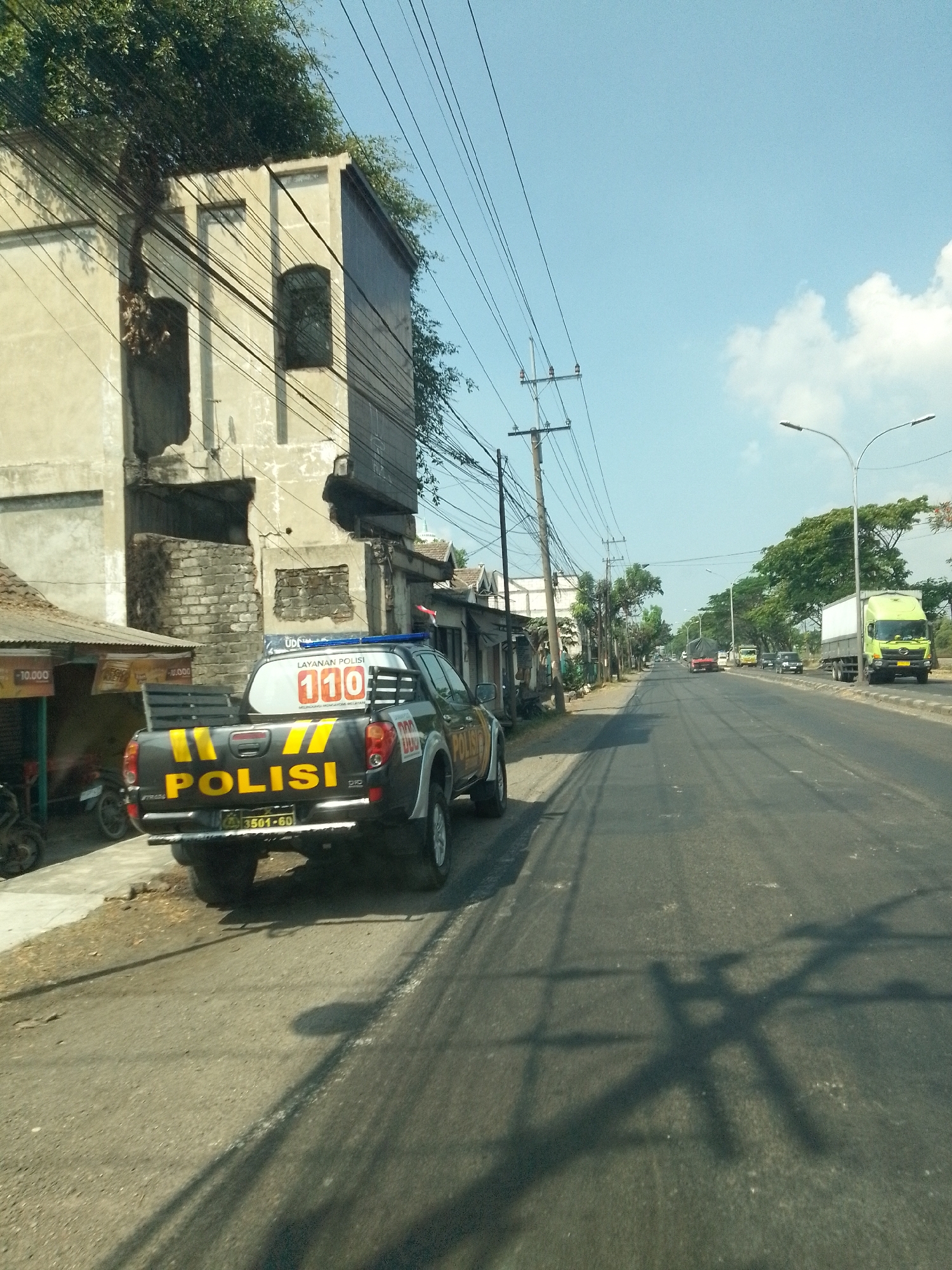 The image captures a street scene in Waruwetan, Indonesia, on a clear, sunny day. A black police pickup truck, marked with yellow stripes and "POLISI" in large letters, is parked on the side of a paved road. The truck has a police light bar on top and a license plate that reads "3501-60". To the left of the truck, a building with light-colored walls and visible structural damage stands. Some small shops with corrugated roofs are also visible to the left. The road is lined with utility poles and power lines, and trees with dense foliage grow along the side. Further down the road, other vehicles, including a green truck, are in motion. The sky is a bright blue with a few scattered white clouds. The overall impression is that of a typical day in a developing urban or semi-urban area, with ongoing activity and infrastructure.