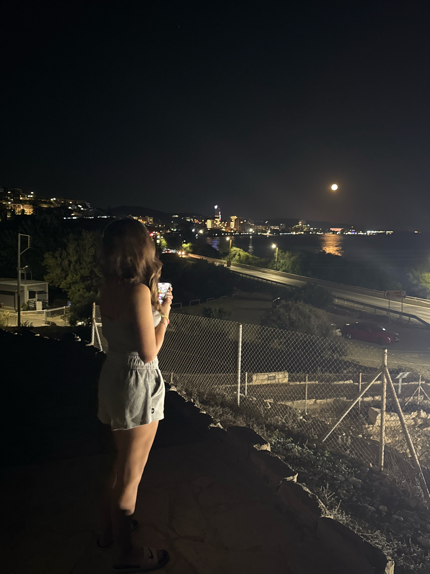 A full-body, side-profile shot captures a young woman standing in the foreground, facing away from the camera, holding a smartphone. She is wearing a light-colored tank top and grey shorts, with sandals on her feet. The background reveals a nighttime cityscape and a body of water, illuminated by numerous lights. The moon is visible in the upper right portion of the sky. A chain-link fence runs across the middle ground, separating the woman from a road and a red car. The terrain in the foreground is rough and uneven, with rocks and sparse vegetation. The setting appears to be Agios Tychona Community, Cyprus, given the distinct view of the coastline and the architecture of the buildings visible in the distance, characteristic of the Limassol coastal area.