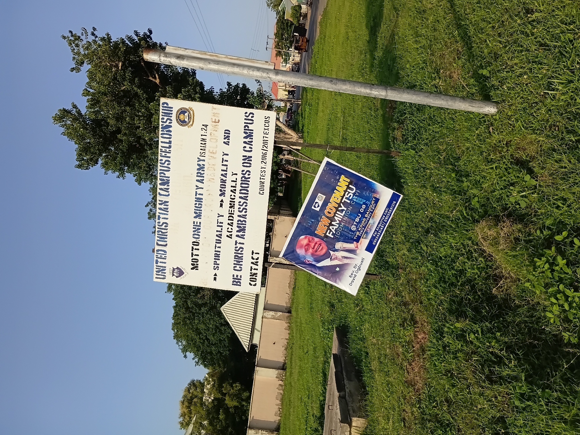 The image shows an outdoor scene in Jalingo, Nigeria, with a large white signpost as the central subject. The signpost has a dark blue emblem at the top and features text in dark blue letters. The text reads: "UNITED CHRISTIAN CAMPUS FELLOWSHIP", "MOTTO: ONE MIGHTY ARMY ISAIAH 1:24", "SPIRITUALITY MORALITY AND ACADEMICALLY", "BE CHRIST AMBASSADORS ON CAMPUS", and "CONTACT". Below this main sign, a smaller poster is attached, displaying a portrait of a Black man in a suit and tie. This poster also has text that is partially visible, including what appears to be "NEW COVENANT FAMILY TSU (DOMINION CITY)" and "REV. DR. DAVID OGBUELI".
The signpost is situated in a grassy area, with a few concrete structures visible in the foreground. In the background, there are buildings, trees, and a road with a three-wheeled vehicle. The sky is clear and blue. The overall impression is that of a college campus or a public gathering space in Jalingo.
