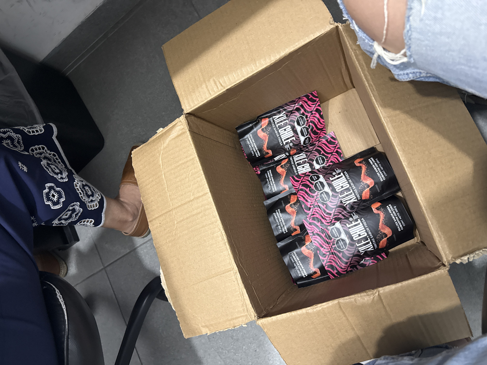 The image depicts a partially filled cardboard box containing six black packages with pink wavy lines and the words "XILE CHILE TIRITAS SABOR FRESA DULCES SABOR FRESA CUBIERTOS CON CHILE CONT. NETO: 200 g." A person wearing ripped denim jeans is seated to the right of the box, and another person wearing blue patterned pants is seated to the left. The setting appears to be indoors, with a tiled floor. The image appears to have been taken from an elevated perspective. The location is San Bernardino Tlaxcalancingo, Mexico, though this cannot be specifically confirmed from the image alone.
