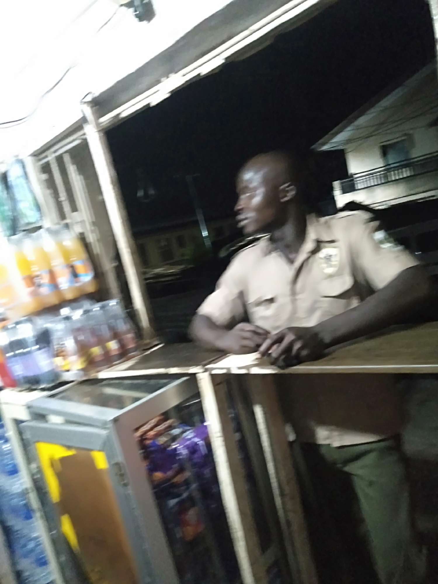 A man in a light brown uniform with dark green pants is standing behind a counter, looking to the right. The counter is part of a small store or kiosk, which contains a refrigerator with drinks and snacks. The store is located in an urban area, with buildings visible in the background. The scene is dimly lit, suggesting it is nighttime. The location appears to be in the city of Unknown, Unknown.