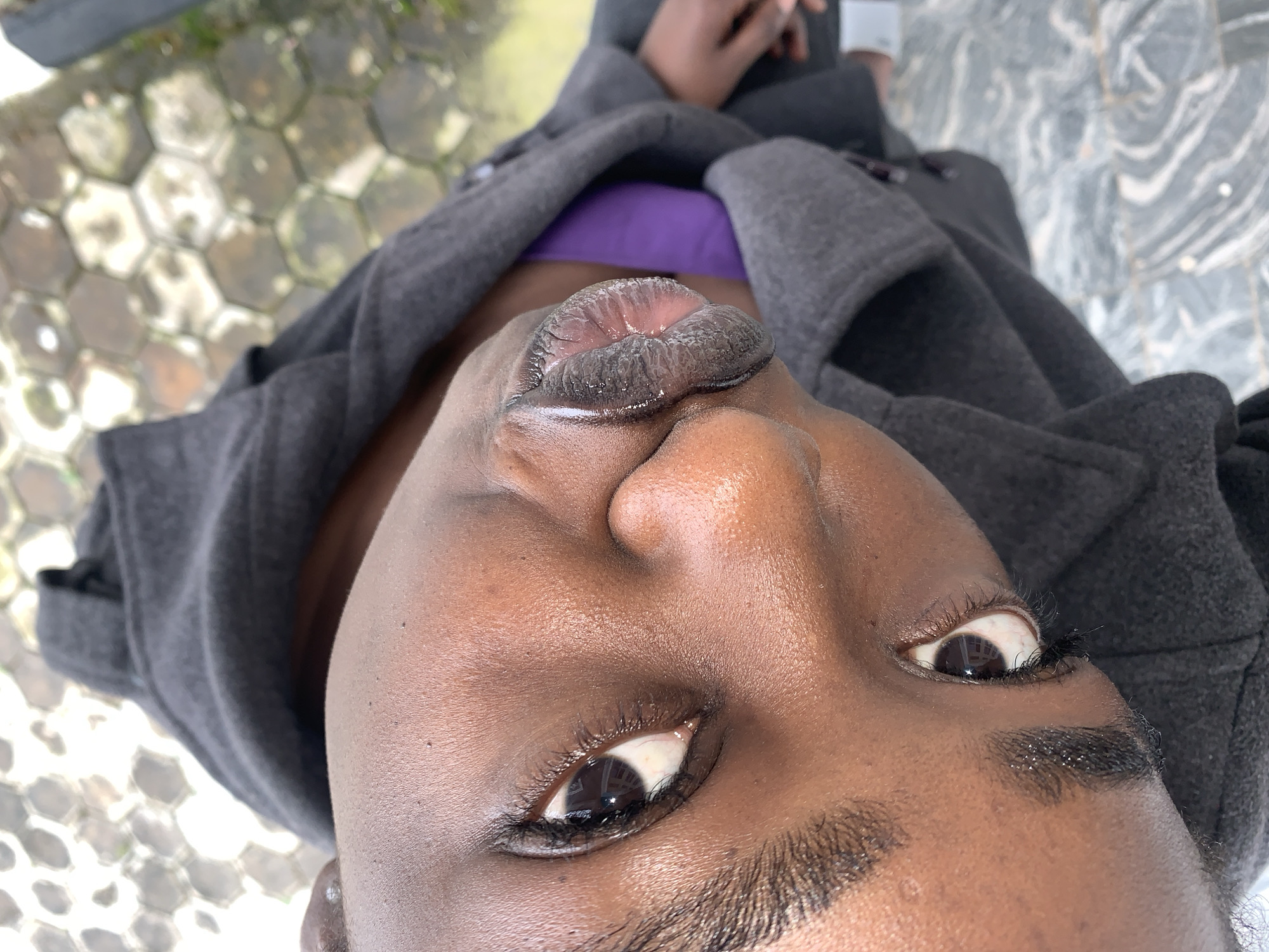 A close-up, head-and-shoulders, slightly angled shot shows a person's face and upper body. The person is wearing a dark gray hooded jacket and a bright purple shirt or garment underneath. The background is out of focus and appears to be an outdoor setting with hexagonal paving stones and some greenery. The person's skin is dark, and their eyes are visible, with one eye more prominent in the frame. Their lips are also a focal point, appearing full and colored a dark shade, possibly lipstick or natural pigmentation, with a textured surface. The lighting suggests it is daytime. The image is oriented such that the top of the person's head is towards the bottom of the frame. The location within Gura Top, Nigeria, cannot be determined from the visual information present in the image.