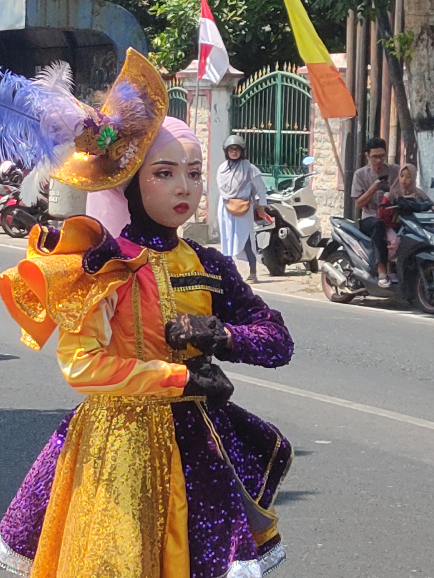 The image captures a vibrant street scene in Ngasem, Indonesia, likely during a procession or festival. The central focus is a person in elaborate ceremonial attire, featuring a headdress adorned with purple feathers and golden, ornate embellishments. Their costume is a dazzling ensemble of bright yellow and deep purple, with shimmering sequins and voluminous ruffled sleeves in orange and gold. The individual is wearing a light pink hijab and dark gloves, and their face is artistically made up with glitter and dramatic eyeliner.
In the background, the environment is distinctly Indonesian. A green, wrought-iron gate suggests a building or compound. Various motorcycles are parked and in motion, indicating a busy street. People are seen on or near the motorcycles, some observing the procession, others engaged in their daily activities. Flags, one predominantly yellow and orange, flutter in the background, adding to the festive atmosphere. The asphalt road and sidewalk are visible, characteristic of a typical urban street in Indonesia. The overall lighting suggests it is a bright, sunny day.