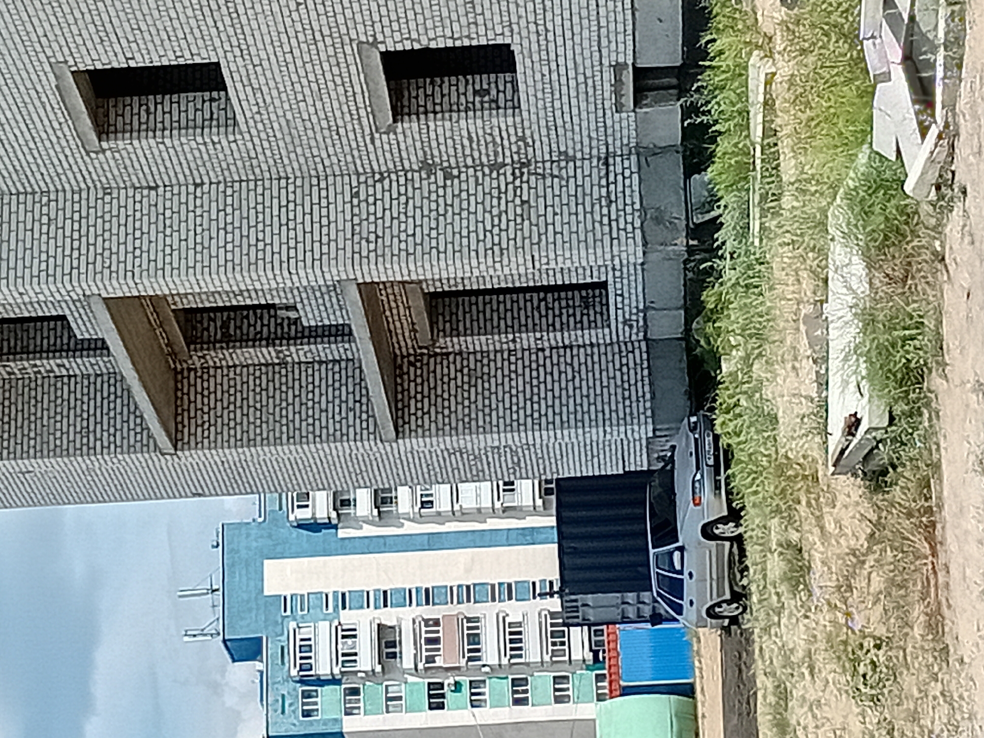 The image depicts a construction site in the city of Semey, Kazakhstan, under a partly cloudy sky. In the foreground, a silver hatchback car, possibly a Lada Samara, is parked on a dirt lot overgrown with tall, dry grass and some construction debris. Behind the car, a dark shipping container is visible, along with a blue corrugated metal structure. Further in the background, there are several multi-story residential buildings. The building on the right is under construction, made of white brick, with unfinished window openings and concrete balconies. The buildings on the left are taller and appear to be older, with blue and white facades and many windows. The overall scene suggests an ongoing urban development project in Semey.