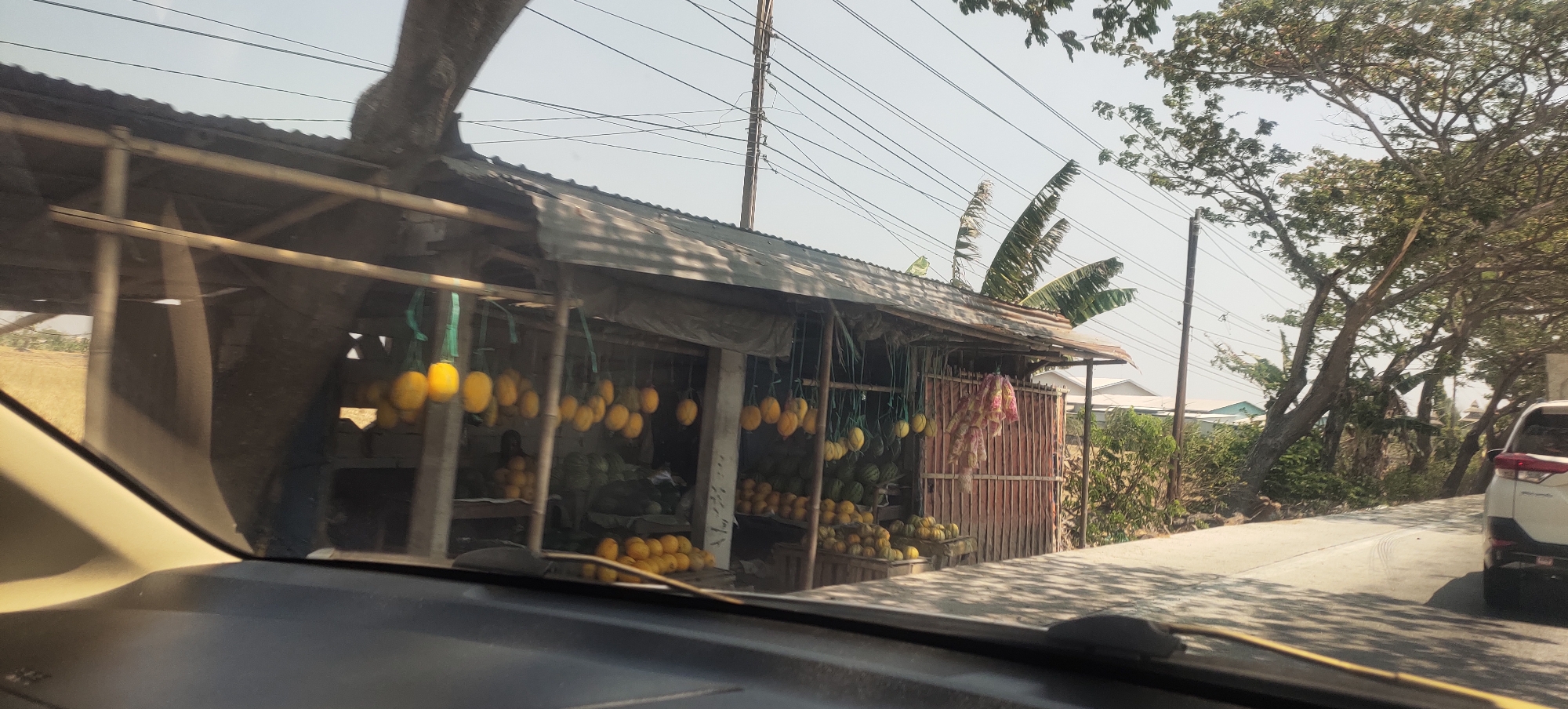 The image depicts a roadside fruit stand in Sedo Lor, Indonesia, viewed from the perspective of a vehicle. The stand features a corrugated metal roof supported by bamboo poles, with a tarp or cloth draped over the front. Numerous yellow fruits, likely mangoes, are hanging from strings. Below the hanging fruits, various other fruits, including what appear to be watermelons and smaller round fruits, are displayed in crates and on the ground. A person is partially visible behind the counter within the stand. In the background, trees, power lines, and a few buildings are visible. A white car is parked on the right side of the road, adjacent to the fruit stand. The scene is bathed in daylight, with clear skies.