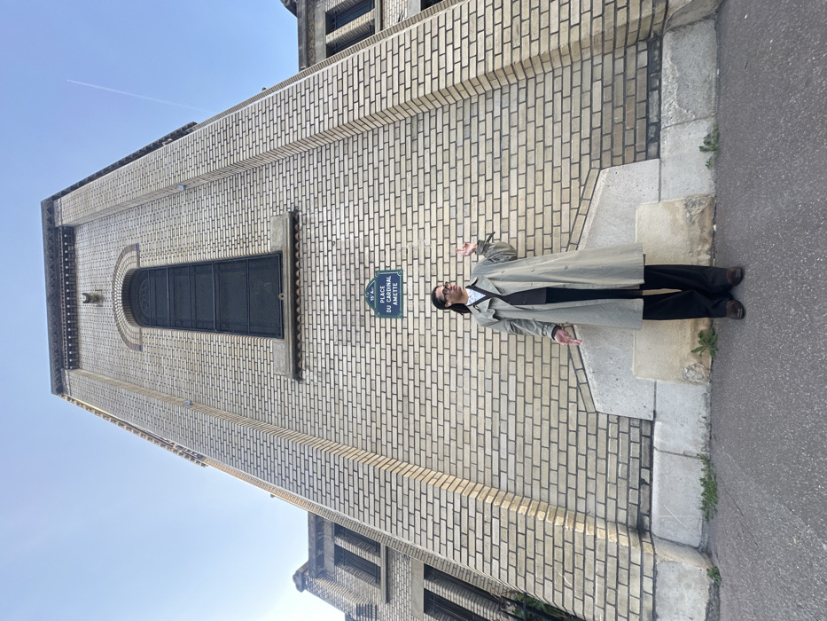 The image displays a person standing outdoors next to a large brick building, under a clear blue sky. The photo is taken during daytime, and a faint airplane contrail is visible in the upper left of the sky.
The central figure is an adult person with dark hair, wearing glasses, a white collared shirt, dark trousers, and a long, light-colored trench coat. They are standing with their arms slightly outstretched and hands open, in a gesture that suggests posing for the camera, perhaps expressing surprise or emphasizing something. The person is looking slightly upwards and to their left.
Behind and to the left of the person is a prominent building constructed from light-colored bricks, laid in a pattern with dark mortar lines. The building has a historical or institutional architectural style, featuring a large, dark-framed arched window with intricate detailing above the person's head. Below this window, a small, dark object or feature protrudes from the wall. Further down the wall, closer to the person's right, is a smaller rectangular, dark-framed window.
Mounted on the brick wall to the right of the person's head is a blue street sign with white text. The text on the sign reads:
"15e Arrt."
"PLACE DU CARDINAL"
"AMETTE"
This indicates the location is the Place du Cardinal Amette in the 15th Arrondissement of Paris, France.
The ground in the foreground consists of a grey sidewalk or curb directly beneath the person's feet, separating the building from what appears to be a paved street surface on the far right. Small patches of green vegetation are visible growing from cracks in the sidewalk. Another part of the building, possibly a lower section or an adjacent structure, is visible in the lower right portion of the image.