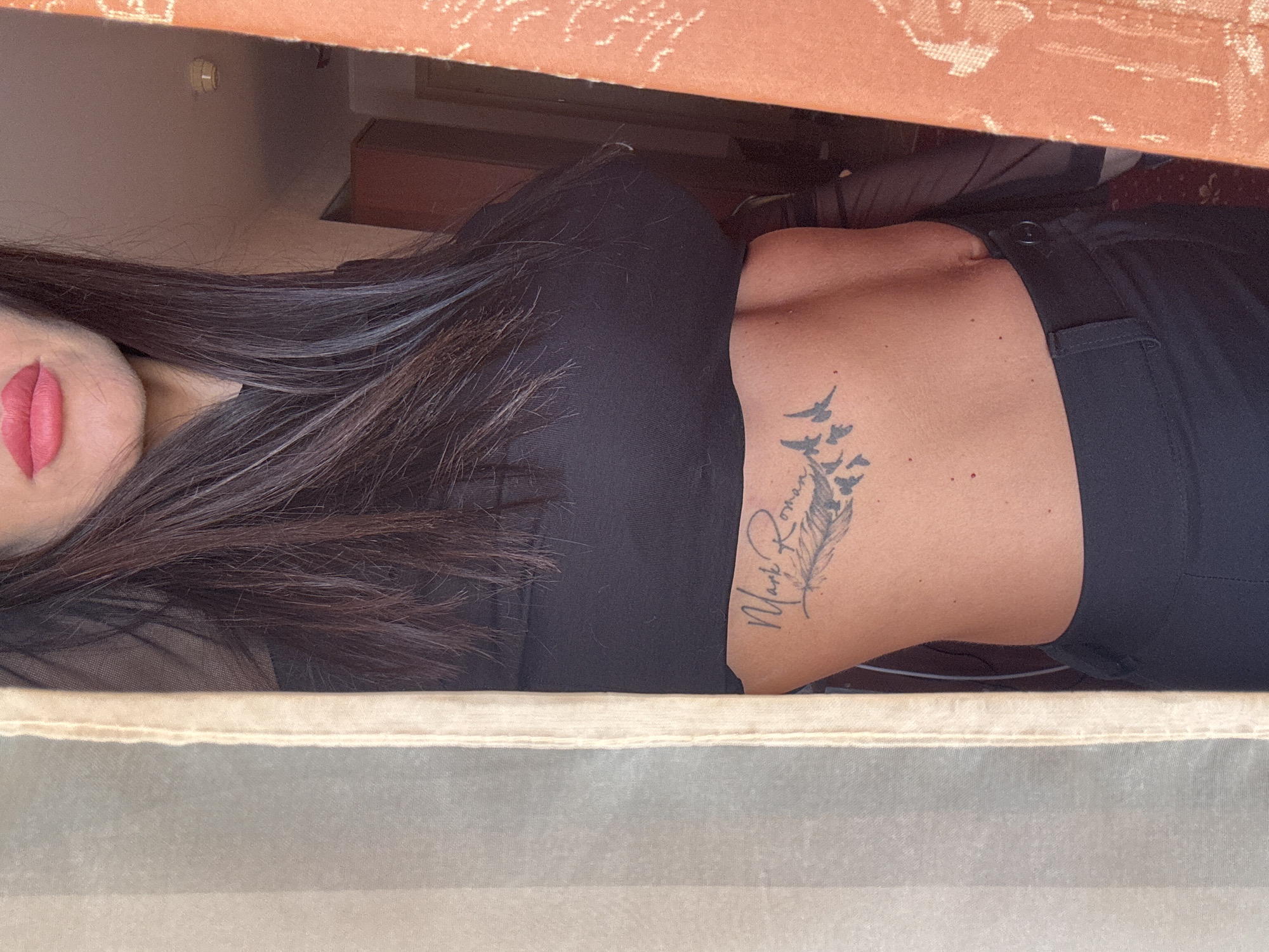 A woman with long, dark hair and red lipstick is seen partially through a frame, likely a window or doorway. She is wearing a black top and black pants, and she has a tattoo on her side featuring a feather and the name "Mark Ronan". The location in the city of Perm, Russia, is not visually verifiable from the image. The setting appears to be indoors.