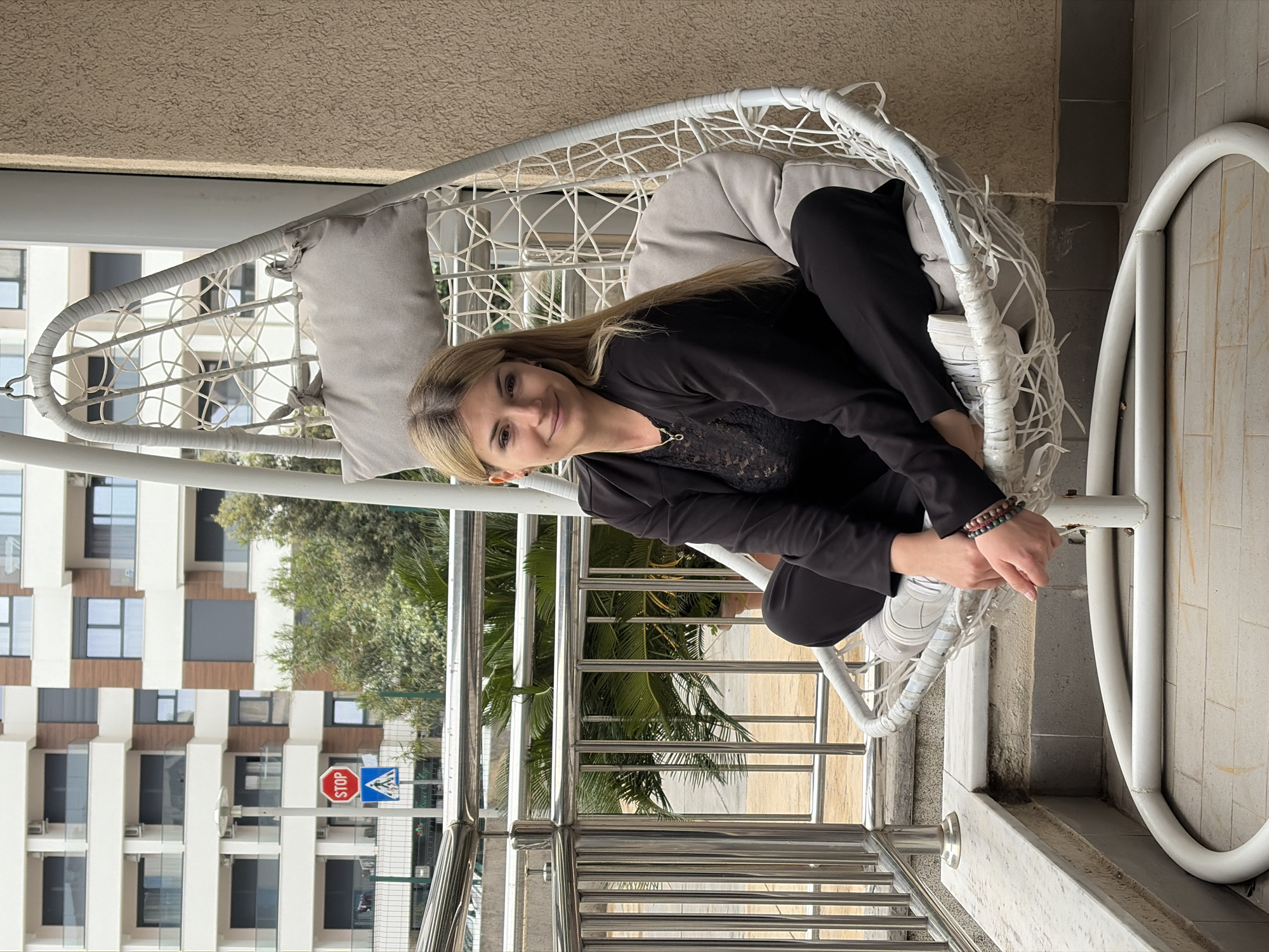 A woman is seated in a white, egg-shaped swing chair on a balcony, with a pleasant urban landscape in the background. She has long, light-colored hair and is wearing a black long-sleeved top with a lace detail, black trousers, and white sneakers. Beaded bracelets adorn her left wrist. She is looking towards the camera with a calm, subtle smile. The swing chair is cushioned with light grey pillows.
The setting appears to be a balcony, complete with a modern metal railing. Beyond the railing, multi-story buildings with large windows and varying facades suggest an urban environment, characteristic of a city like Budva, Montenegro. Lush green trees and palm-like foliage are visible between the buildings, adding a natural element to the scene. Below, a street or road is visible, featuring a prominent red octagonal "STOP" sign and a blue triangular warning sign. The clear, natural lighting indicates it is daytime with good weather. The overall atmosphere is relaxed and serene.