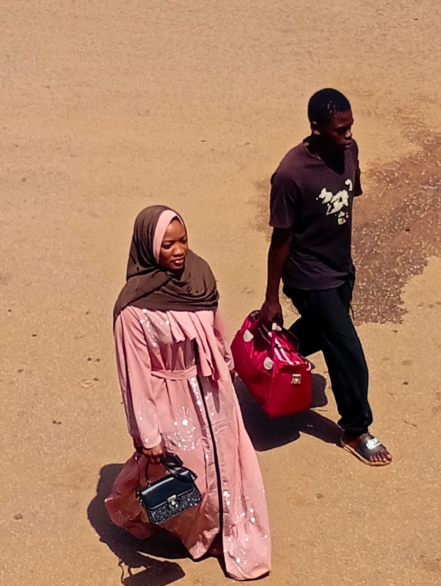 In Bukuru, Nigeria, a man and a woman are walking across a sandy surface. The woman, on the left, is wearing a pink abaya and a dark headscarf. She is carrying a small black handbag. The man, on the right, is wearing a dark t-shirt with a white graphic and dark pants. He is carrying a bright pink duffel bag in his right hand and is wearing sandals. The ground appears to be dirt or sand, with some patches of wetness or darker discoloration. The scene is captured from an elevated, overhead perspective.