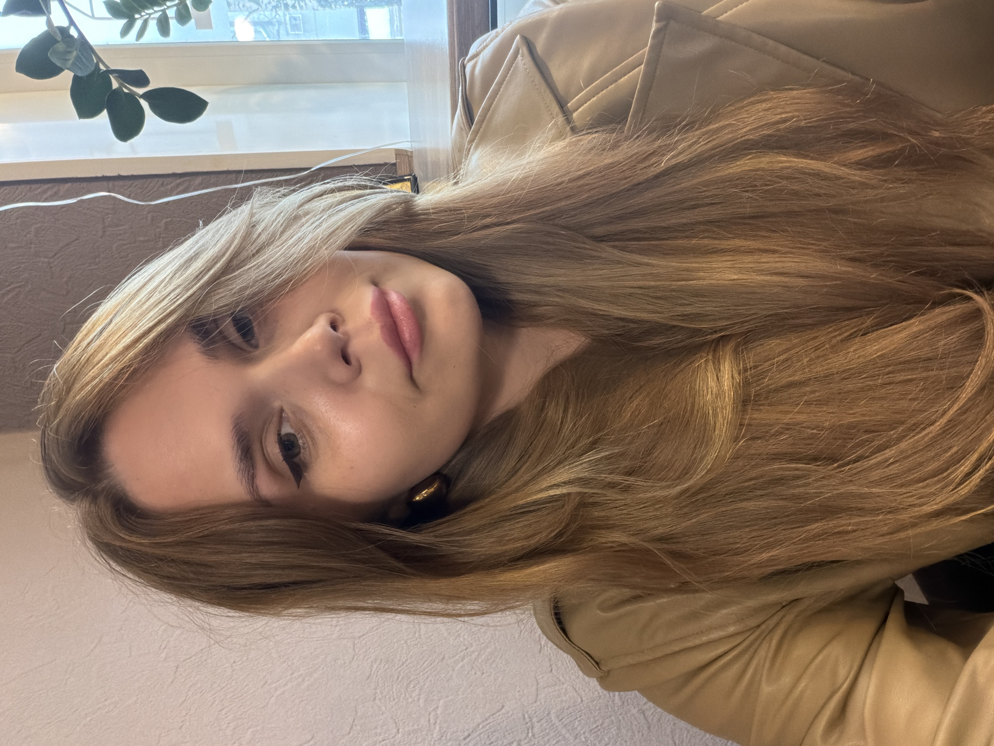A young woman with long, light brown hair is posing for a selfie. She is wearing a light brown leather jacket. A window with plants in front of it is in the background. The location is Chaikovsky, Russia.