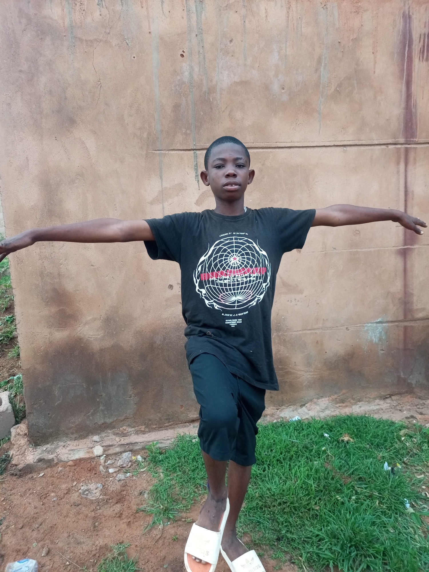 A young boy stands in front of a textured, tan-colored wall, his arms outstretched horizontally. He is wearing a black t-shirt with a circular graphic design and black shorts. The boy is balancing on his left leg, with his right leg slightly bent. He is wearing white sandals. The ground in front of the wall is a mix of dirt and patches of green grass. Debris, including plastic wrappers and rocks, is scattered on the ground. The wall appears to be an exterior surface of a building, possibly in an outdoor urban setting in Tempure, Nigeria. The lighting suggests it is daytime.