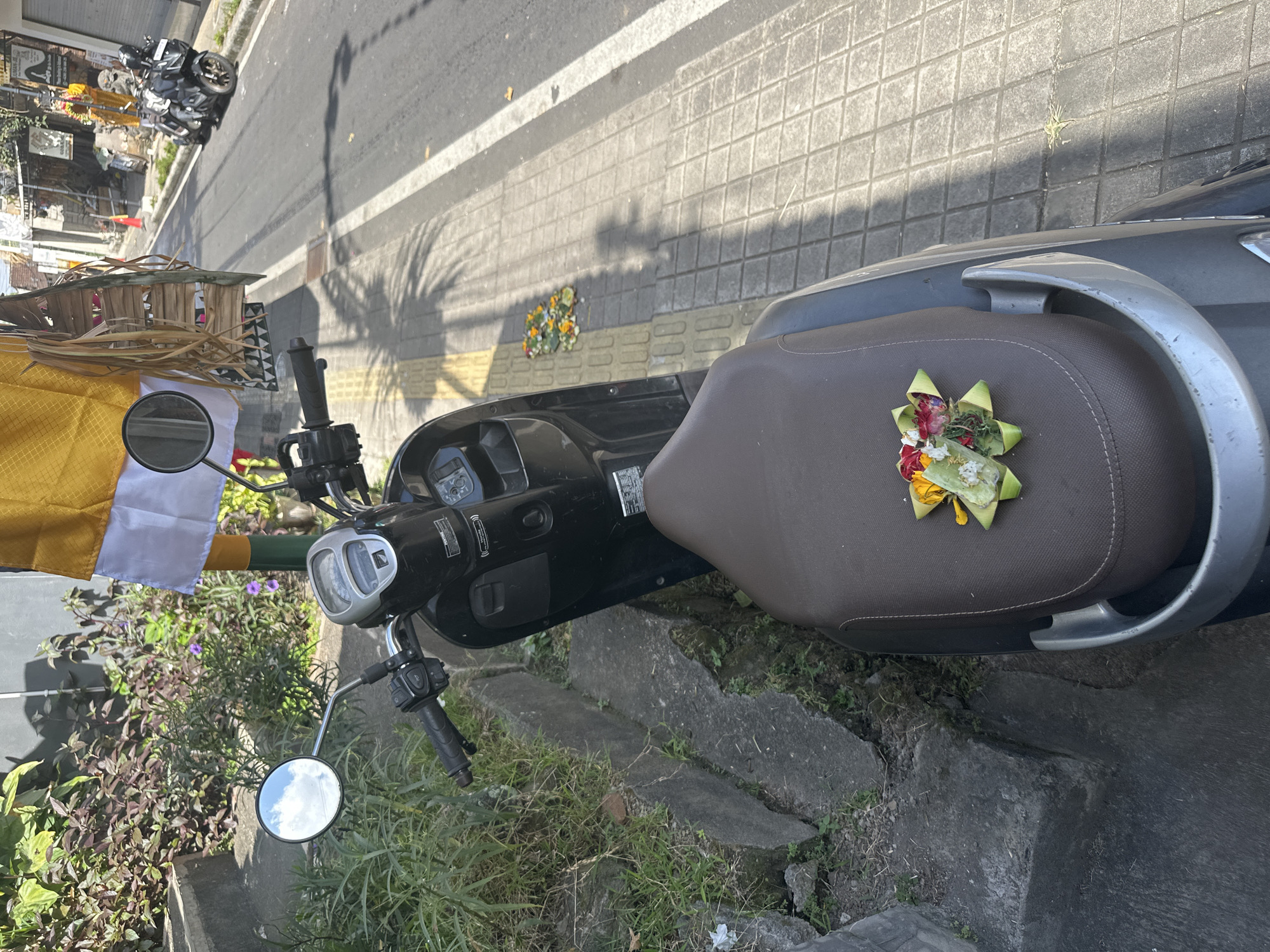 A dark grey scooter is parked on the side of a street in Ubud, Indonesia. The scooter's seat is brown and has a small arrangement of flowers and leaves on it. To the left of the scooter, there is a sidewalk and some green plants. To the right of the scooter, the street is visible, with other scooters parked further down. A yellow and white flag is hanging from a pole to the right of the scooter. The scene appears to be in an urban environment, possibly a residential or commercial area. The image is taken from a high angle, looking down at the scooter and the surrounding area.