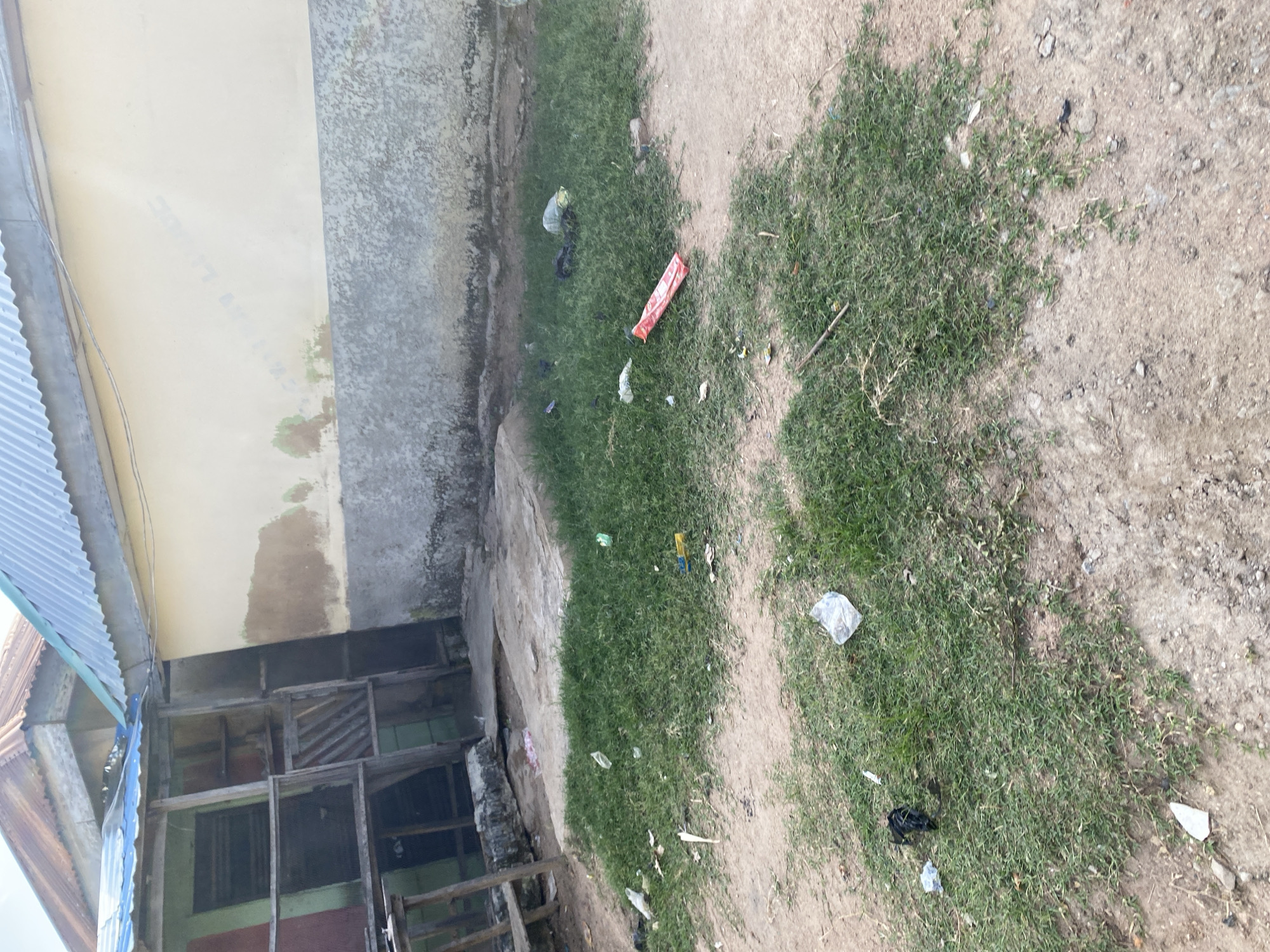 The image depicts an outdoor scene in Jauro Yinu, Nigeria, likely a residential or communal area. The foreground is dominated by patches of green grass interspersed with sandy ground, showing signs of litter. Several pieces of trash are scattered across the grass and ground, including plastic bags, wrappers, and a red plastic object. In the background, a building with a light-colored wall and a weathered, textured lower section is visible. Adjacent to this building is a smaller structure with a corrugated metal roof, and an open-sided, somewhat dilapidated wooden structure with multiple compartments and shelving. The overall impression is one of an unkempt and possibly impoverished environment.