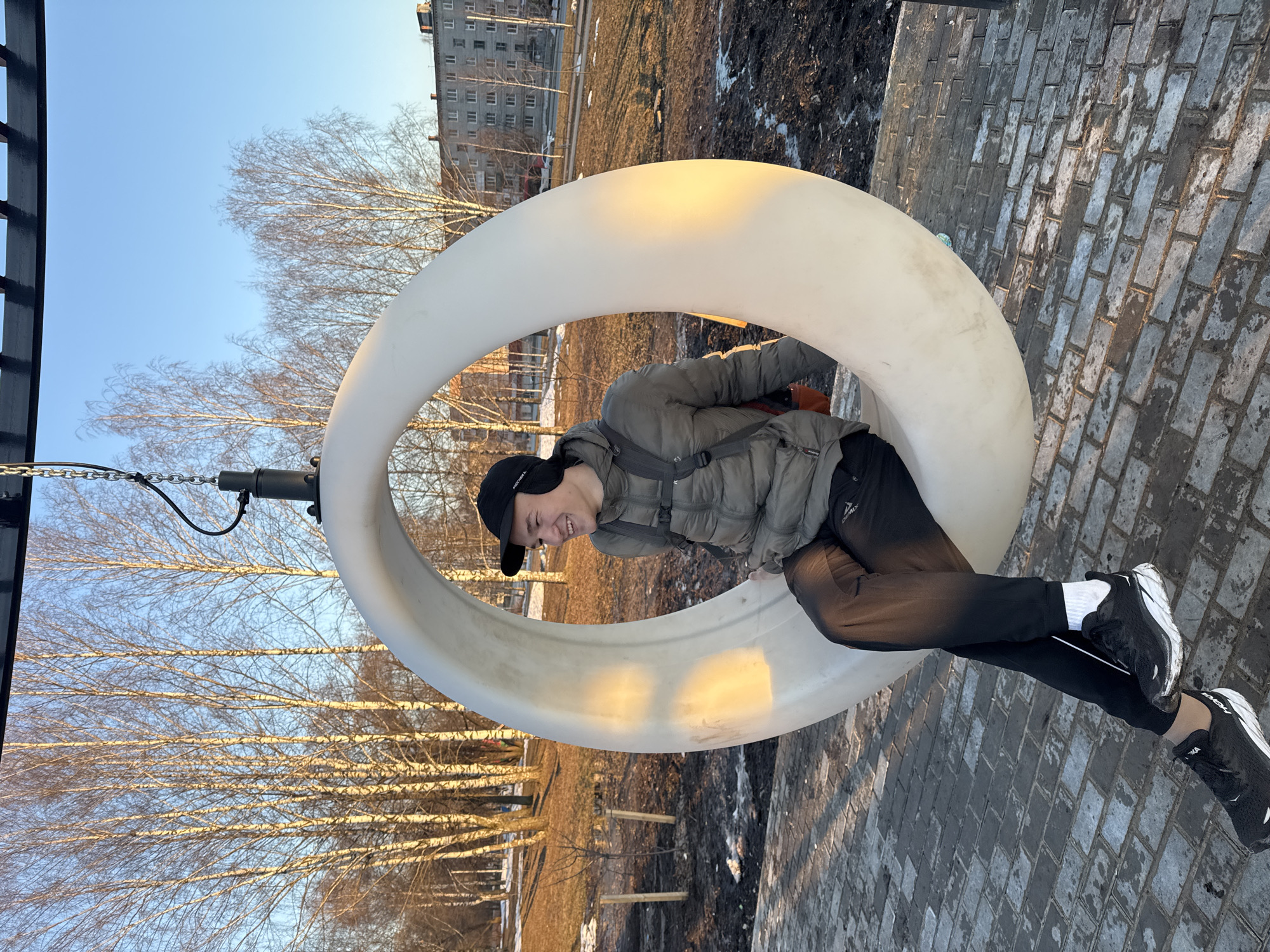 The photograph shows a young man on a tire swing in a park in Berezniki, Russia. He wears a black baseball cap, a gray puffer jacket, black pants, and black and white sneakers. He is smiling. The tire swing is white and hangs from a black metal frame and chains. The ground is paved with gray bricks. Bare trees are visible in the background, along with a multi-story apartment building. There are patches of dirt and remnants of snow in the background. The sky is blue.