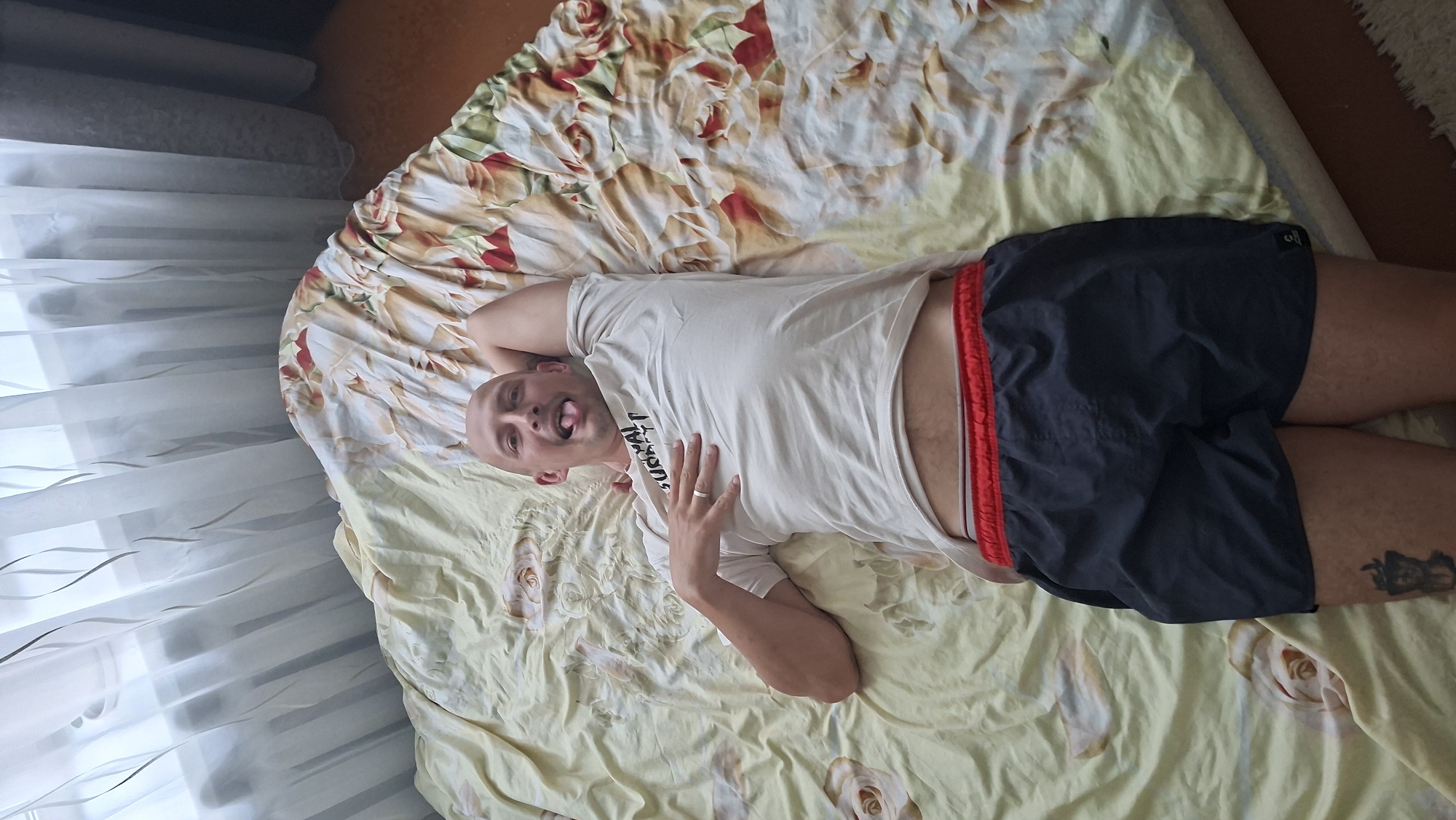 A bald man is lying on his back on a bed with a floral patterned comforter. He is wearing a white t-shirt and dark blue shorts with a red waistband. His left arm is bent behind his head, and his right hand is on his chest. He is sticking his tongue out and looking at the camera. There is a tattoo visible on his left thigh. The background shows sheer curtains and a window. The scene appears to be indoors, possibly in a bedroom. There are no other people or animals present in the image. The overall lighting suggests it is daytime. The location is identified as Myadzyel, Belarus.