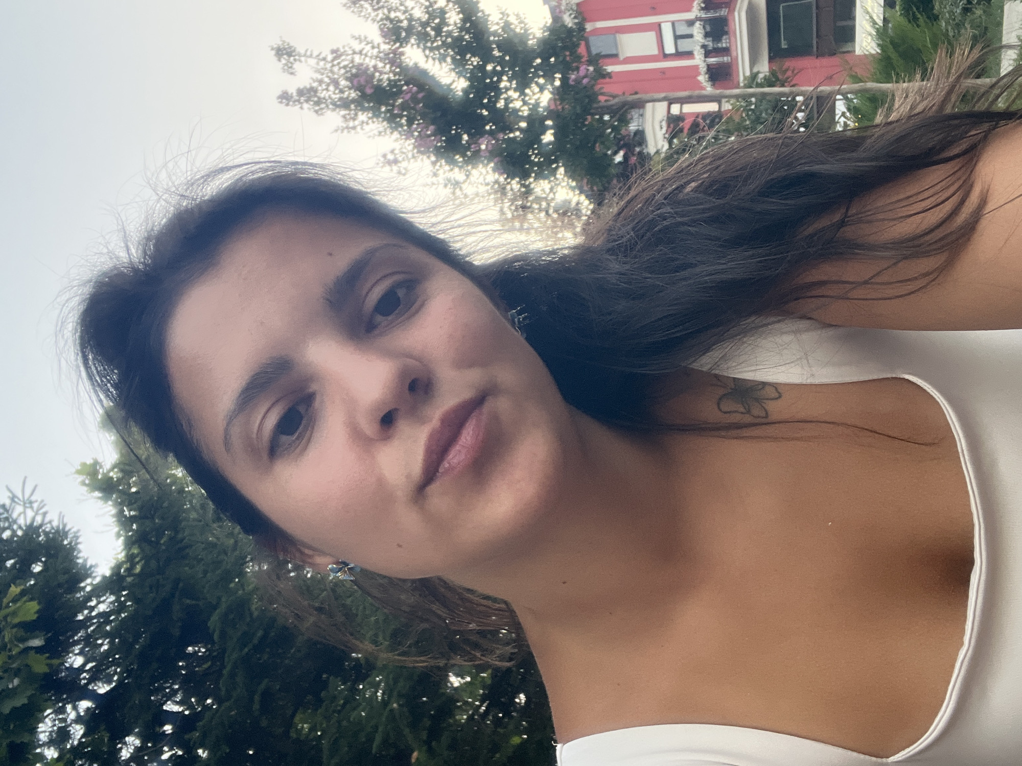 A close-up, selfie-style photograph shows a young woman with dark hair, looking directly at the camera with a neutral expression. She is wearing a white top and a small butterfly tattoo is visible on her left shoulder blade. The background features greenery and a red building, suggesting an outdoor urban setting. The lighting indicates it is daytime, possibly late afternoon. The scene is consistent with a residential or commercial area in the city of Plovdiv, Bulgaria, given the architectural style of the building and the surrounding vegetation.