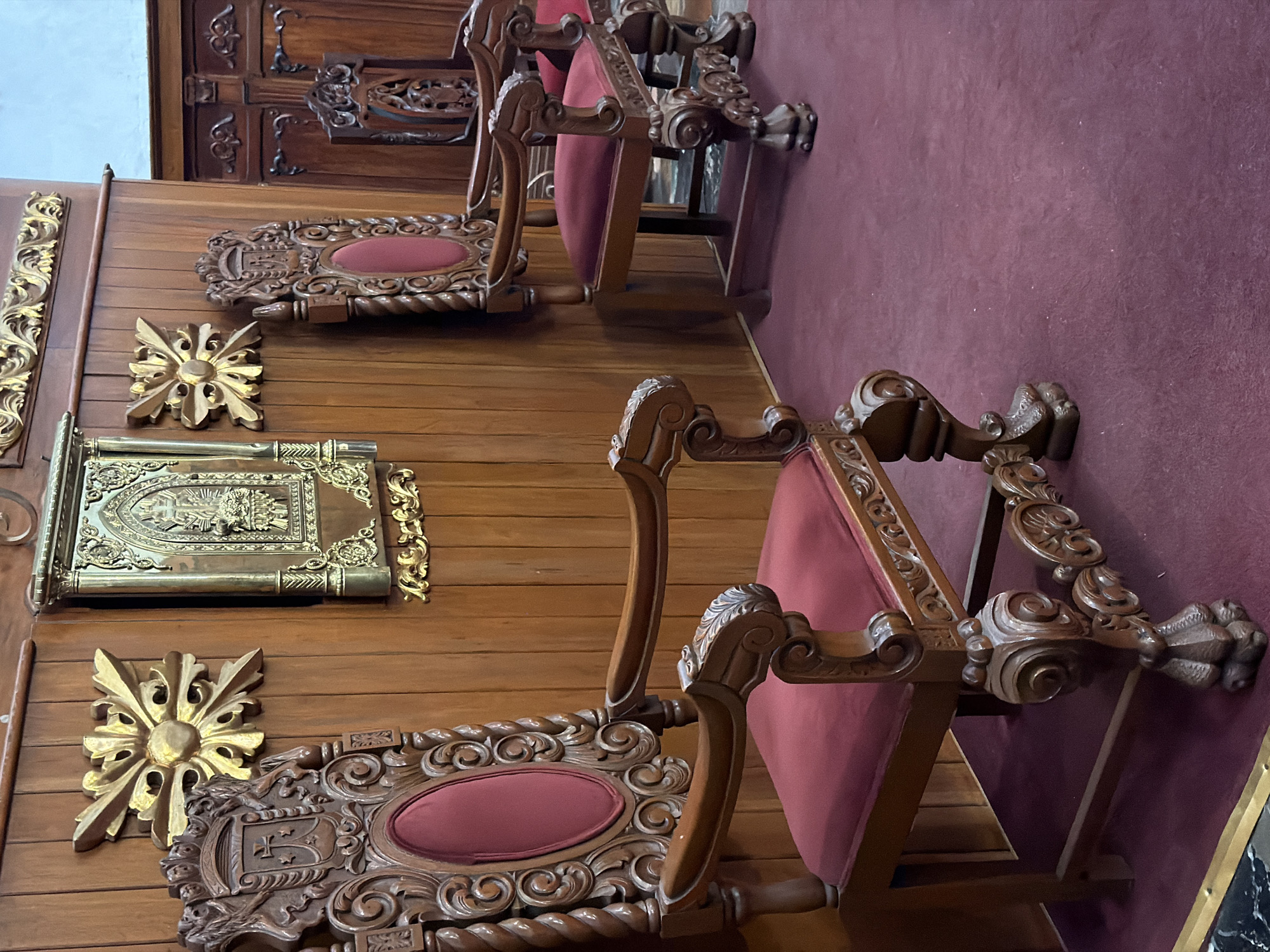 The image shows a section of a room with ornate, dark wood furnishings. Two chairs with red cushions and elaborate carvings are prominent. One is closer to the viewer, showing the intricate details of its frame. Behind the chairs, a wooden wall features decorative gold elements, including floral-shaped appliques and a framed object with gold accents. A burgundy carpet is visible to the side of the chairs. The setting appears to be an old and ornate interior, potentially a church or a historical building. The location is Unknown, Unknown.