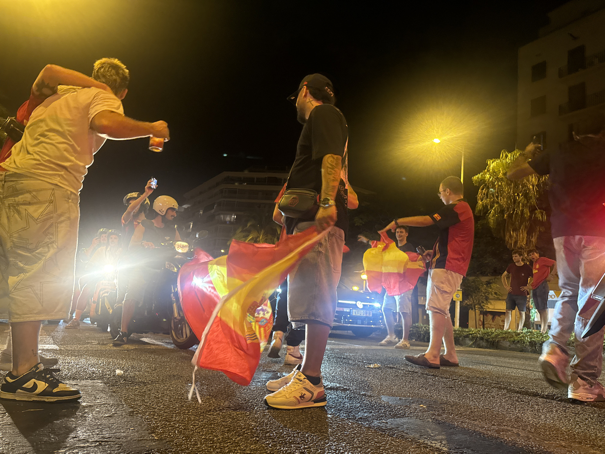 At night in Barcelona, Spain, a group of people are celebrating in the street. A person in the foreground is holding up a Spanish flag, which is red and yellow. The flag is partially obscuring the person's legs and feet. They are wearing white shorts and black and white sneakers. To the left, another person is holding a drink and looking to the right. They are wearing a white t-shirt and light-colored shorts with a star pattern. In the background, several motorcycles are parked and people are gathered around them. One person on a motorcycle is wearing a helmet and has their arms raised. There are buildings visible in the background, and streetlights are illuminating the scene with a warm, yellow glow. The street appears wet, suggesting it may have rained or been recently cleaned. Several other people are seen in the background, some holding flags, others standing and observing. One person in the mid-ground is wearing a black t-shirt and a black baseball cap, and has a fanny pack around their waist. Another person further back is wearing a red t-shirt and light-colored shorts. The overall atmosphere appears festive and energetic.