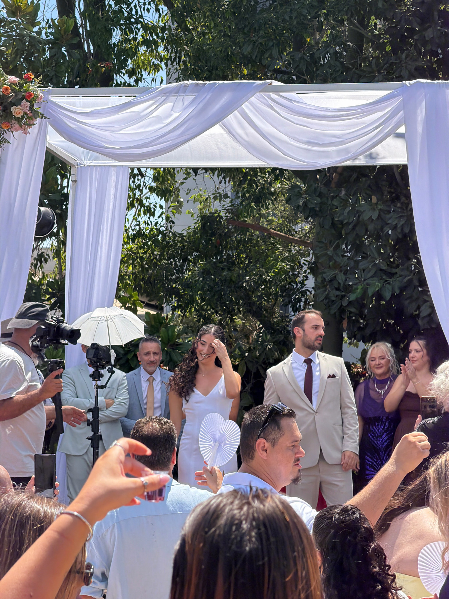 In Kadima, Israel, a wedding ceremony is taking place outdoors under a white draped canopy adorned with flowers on the left side. A man in a light-colored suit with a maroon tie and pocket square stands to the right of a woman in a white wedding dress. The woman is smiling and touching her hair. Behind them, several guests are visible. To the left, a cameraman wearing a hat is filming the event. Other individuals are holding cell phones and fans. The environment is characterized by lush green trees and sunlight filtering through the foliage.
