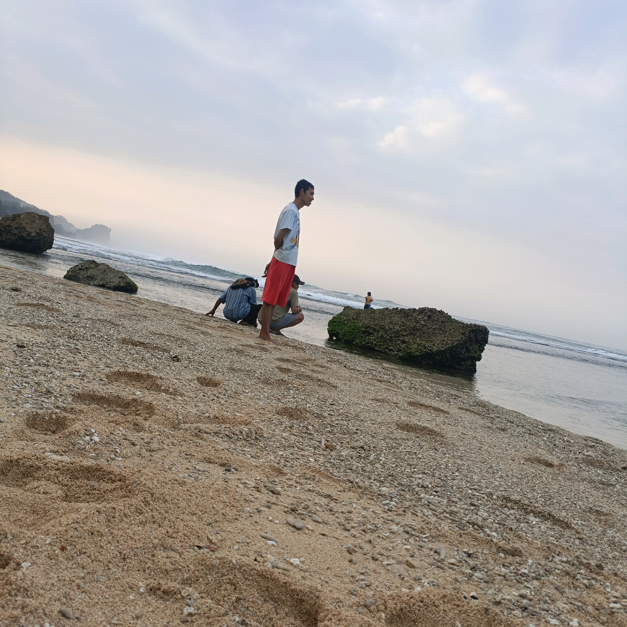 The image shows a beach scene on the coast of Tepus, Indonesia, under a cloudy sky. In the foreground, a sandy beach is covered with small pebbles and shells, with footprints visible. Three individuals are present on the beach. One man in a light-colored t-shirt and red shorts stands facing the ocean, his hands behind his back. To his left, two other individuals are crouched down near the water's edge. The person on the left is wearing a striped shirt, and the person on the right is wearing a light-colored t-shirt and dark shorts. Further down the beach, a large rock covered in moss sits by the water, and another, smaller person is visible in the distance. The ocean is calm with small waves breaking on the shore. In the background, distant cliffs and rock formations are visible through a hazy atmosphere.