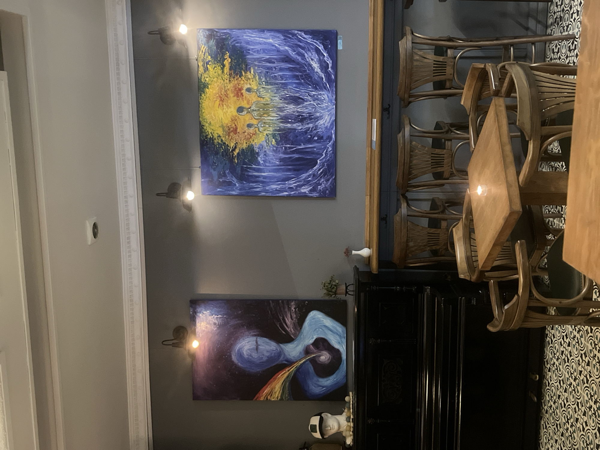 A dimly lit interior space. The left side of the frame features a gray wall with white molding. Two paintings hang on the wall, illuminated by small, directional spotlights. The upper painting is abstract with swirls of blue and a burst of yellow. The lower painting depicts an abstract form in blues, purples, and black, with a colorful stream emanating from it.
To the right, a dark-stained wooden bar and bar stools are visible. A black cabinet or furniture piece is positioned between the paintings and the bar area. A collection of decorative items sits atop the cabinet. Further to the right, a wooden table with chairs is seen against a black-and-white patterned wall. The location is presumed to be in the city of Sofia, Bulgaria, based on the request but is not visually confirmed.