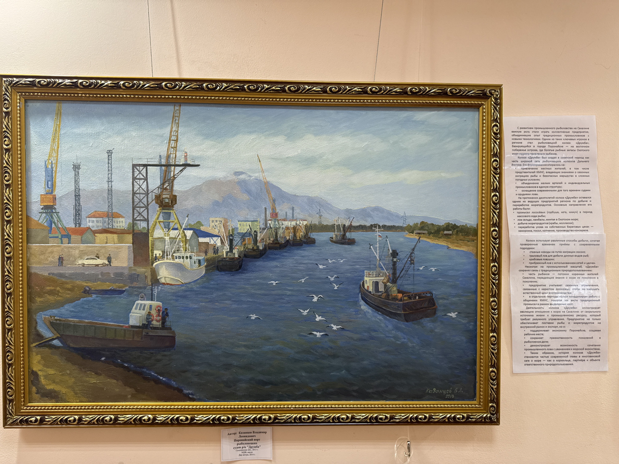 The painting depicts a bustling harbor scene in Poronaysk, Russia, a city located on Sakhalin Island. The foreground features a calm, blue body of water reflecting the overcast sky. Several fishing vessels are anchored or moving within the harbor. On the right side of the water, a dark-hulled fishing boat with a white cabin is prominent, with seagulls flying around it. In the left foreground, a smaller, flat-bottomed boat with three people onboard is pulled up onto a sandy bank.
The midground is dominated by the harbor's infrastructure. A line of larger fishing boats is moored along a quay. Cranes of various sizes and designs are positioned on the shore, suggesting activity related to loading and unloading. Buildings, some with red roofs, are visible behind the quay, and in the distance, mountains form the backdrop. The sky is a pale blue with scattered clouds.
The painting is framed by an ornate gold frame. A text plaque is affixed to the bottom left of the frame, and another text plaque is to the right of the painting. The plaque on the left reads: "Автор: Казанцев Владимир Леонидович. Поронайский порт рыбацких судов р/к "Дружба". Сахалинская область, 2013 г. Масло, холст. Для автора, 2014 г." This indicates the artwork is titled "Poronaysk Port of Fishing Vessels r/k 'Druzhba'", created by Vladimir Leonidovich Kazantsev in 2013, and is for the author's personal collection, dated 2014. The text on the right provides historical context about the development of commercial fishing in Poronaysk and the collective farm "Druzhba".