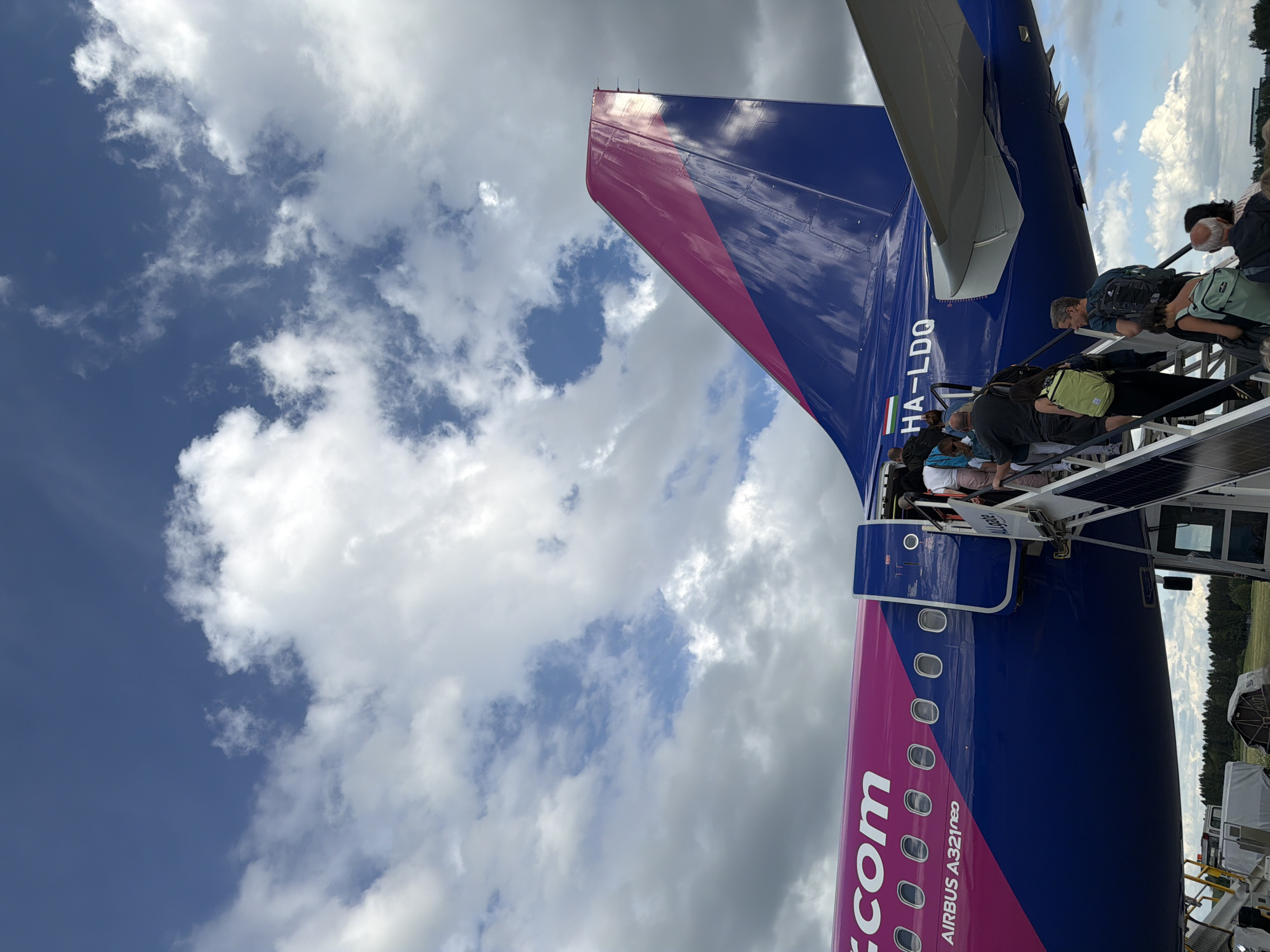 An aerial view of the tail of a Wizz Air Airbus A321neo airplane, registered as HA-LDQ, against a cloudy blue sky. Passengers are boarding the aircraft via stairs on the right side of the image. The airplane's fuselage is predominantly blue with a pink stripe and the Wizz Air logo visible. The Hungarian flag is also visible on the tail. The scene appears to be at an airport in Memmingen, Germany, with a tree line in the background.