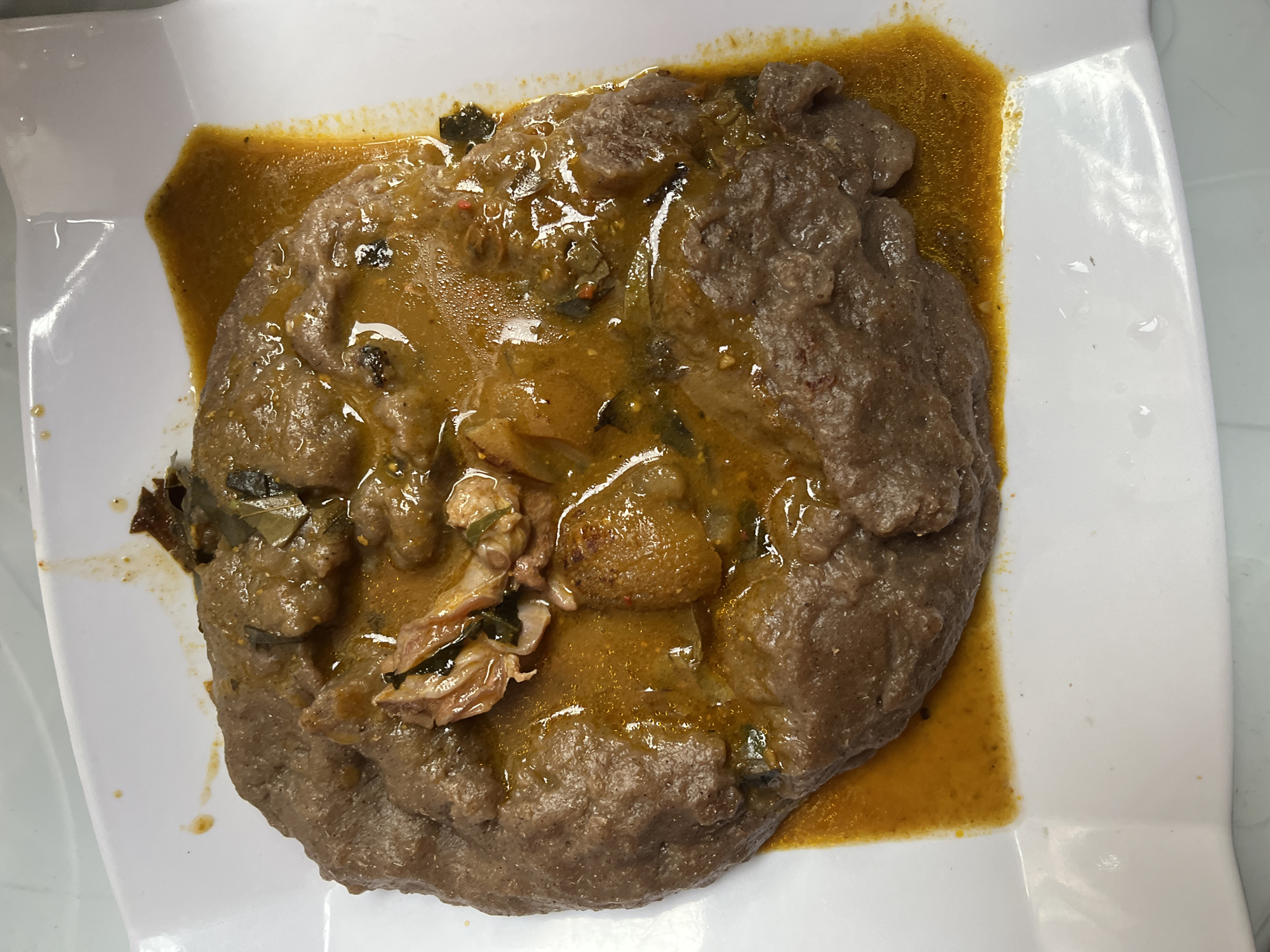 An overhead shot presents a hearty meal served on a pristine white, rectangular plate with slightly raised edges. The central focus is a large, dark-brown, dense mound of a starchy staple food, characteristic of a "swallow" like Amala or Pounded Yam common in West African cuisine. This staple food is partially submerged in, and surrounded by, a rich, glossy, reddish-orange stew or soup.
The stew is glistening with oil and appears to be well-seasoned. Within the stew, various ingredients are visible: chunks of cooked meat, some of which appear to be on the bone, suggesting cuts like goat meat or beef. There are also scattered pieces of dark green leafy vegetables, possibly spinach or scent leaves, contributing to the texture and color of the soup. Some smaller, lighter-colored, rounded pieces are also interspersed within the stew.
The plate itself is clean, with minimal splatters of the stew around the edges, indicating a freshly served meal. The lighting is bright and even, highlighting the textures and colors of the food. The scene depicts a prepared dish ready for consumption, likely an indoor setting. Given the provided context of Iranla, Nigeria, this dish is a quintessential representation of Nigerian culinary tradition. No people or visible text are present in the image.