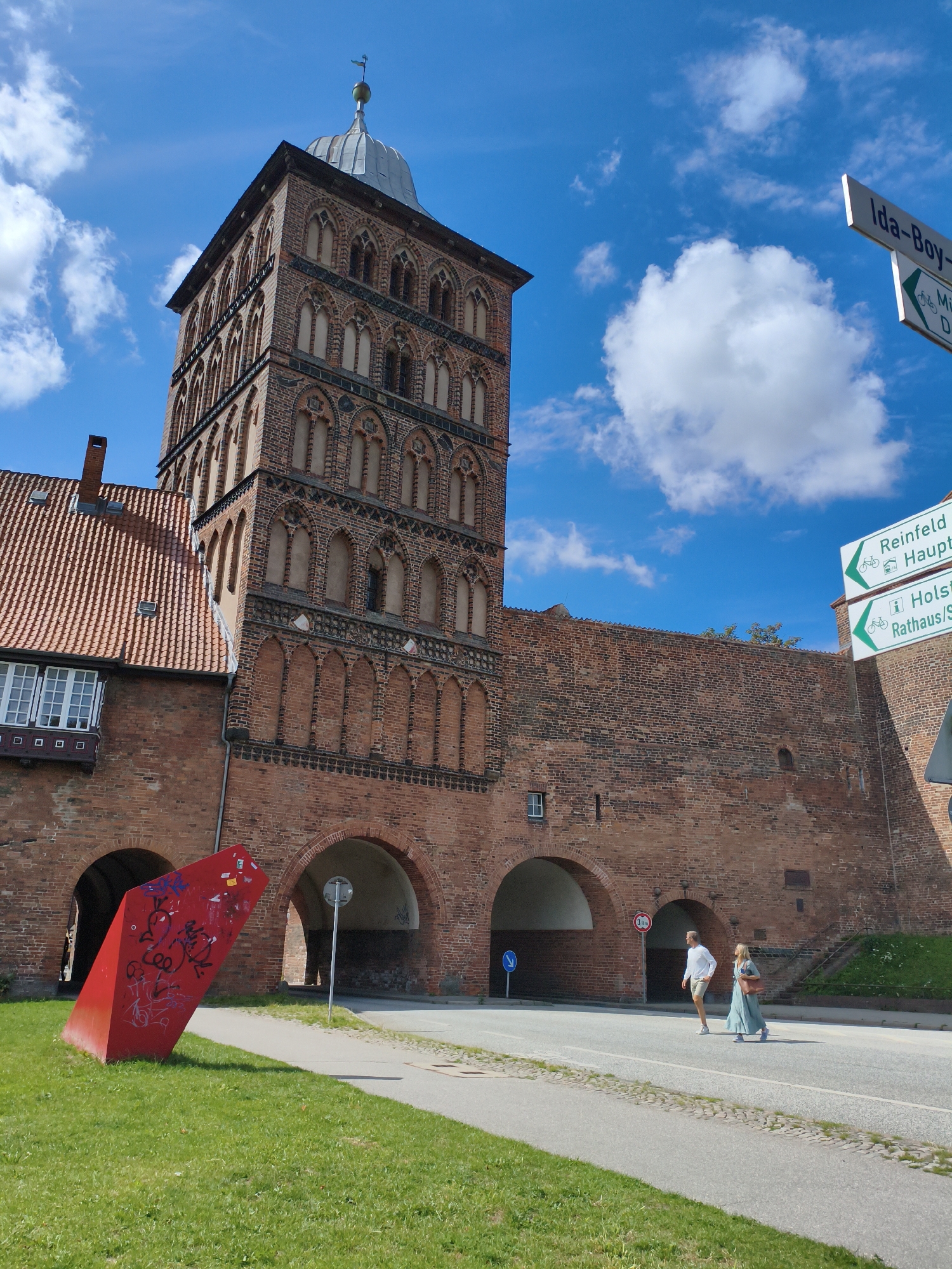 The image depicts the Holsten Gate (Holstentor) in Lübeck, Germany, a historic medieval city gate. In the foreground, a bright red, angular sculpture adorned with graffiti sits on a well-maintained green lawn. To the right of the sculpture, a paved pathway leads towards the imposing brick structure of the Holsten Gate. The gate itself features arched entrances and a tall, narrow tower with intricate brickwork and arched windows. A section of the red-tiled roof of an adjacent building is visible to the left of the tower.
A paved road runs in front of the Holsten Gate. Two individuals are crossing the road; a man in a white shirt and beige shorts is walking, and a woman in a long, light blue dress and carrying a brown handbag is walking beside him. Behind them, several traffic signs are visible, including directional signs pointing towards "Ida-Boy-Str", "Reinfeld Haupt", and "Holst. Rathaus/Str.". A circular red traffic sign with "30" is also present. The sky above is a vibrant blue, dotted with fluffy white clouds. The overall scene suggests a pleasant day in a historically significant urban setting.