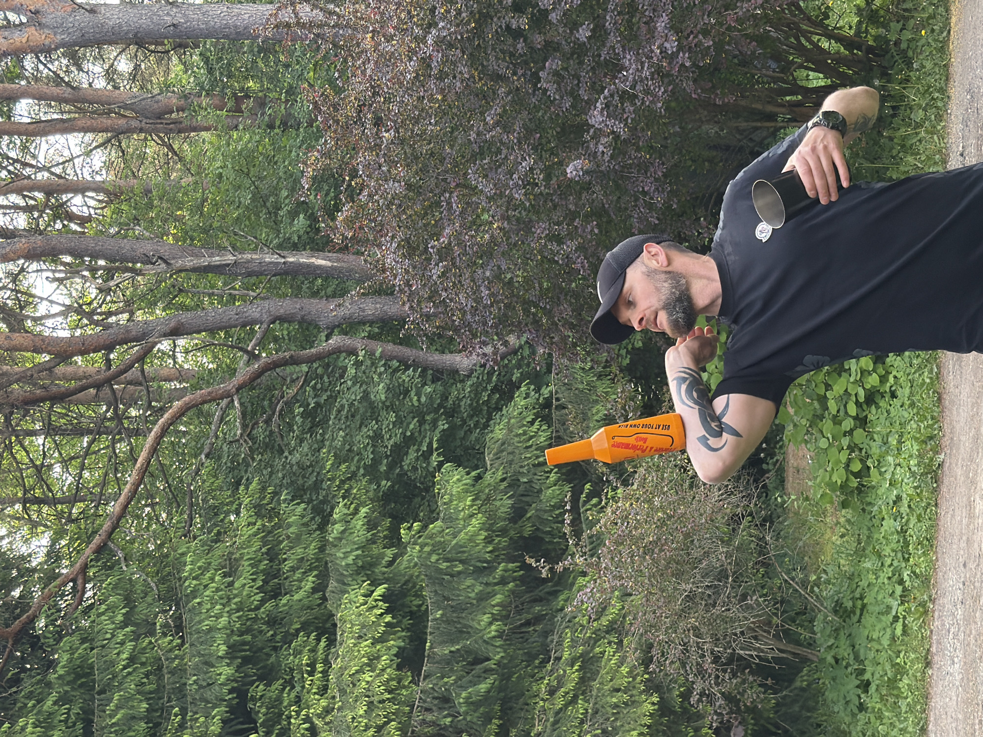 An adult male with a beard and a black baseball cap is outdoors, holding an orange bottle in his right hand and a metal cup in his left. He is wearing a black t-shirt and a watch. He has tattoos on his left arm. The background consists of trees and foliage, suggesting a park or wooded area. The image does not provide enough information to determine a specific location in Tallinn, Estonia.