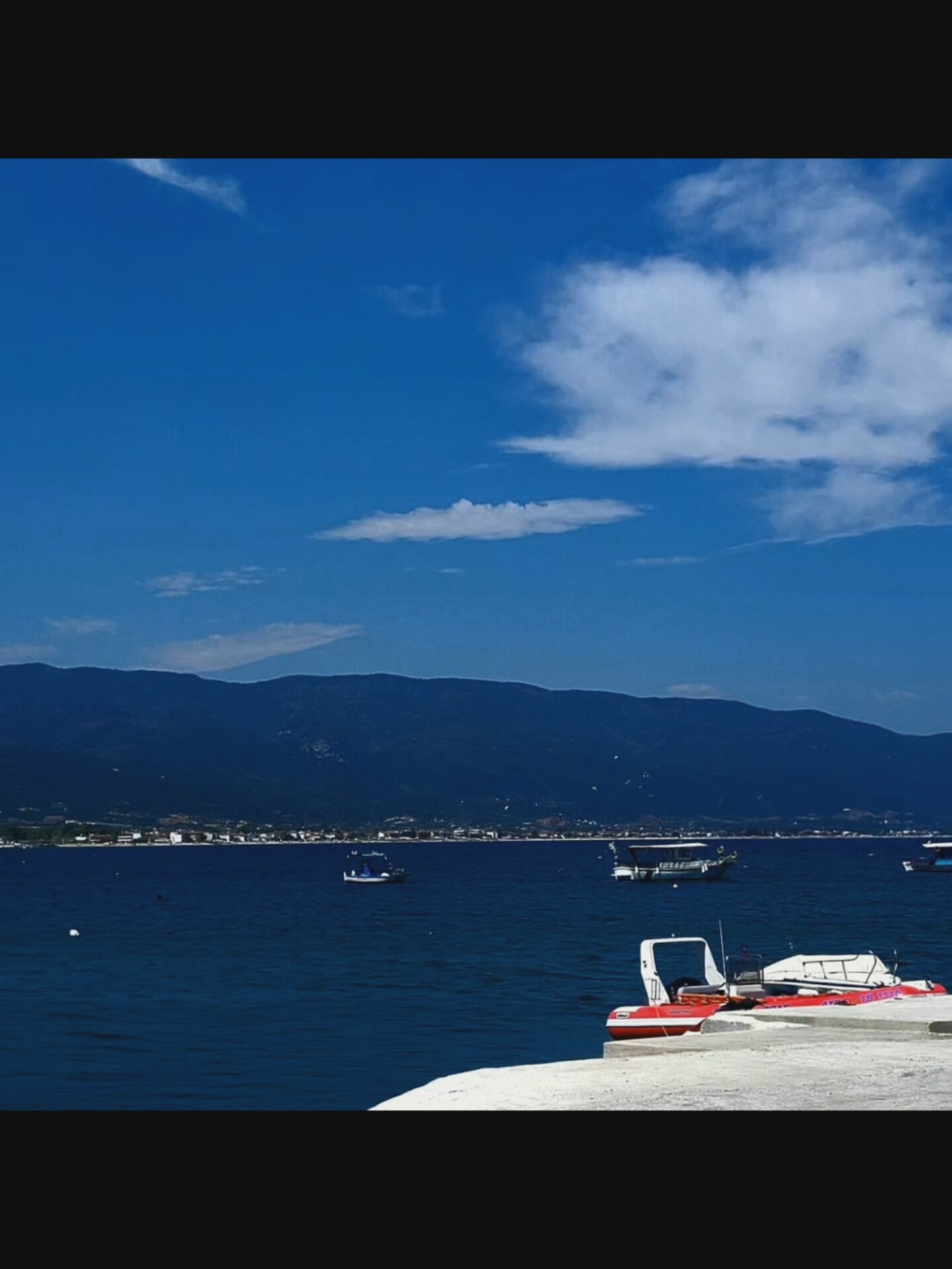 The image depicts a serene seascape with a calm, deep blue ocean stretching towards a distant, dark blue mountain range. The sky above is a vibrant blue, punctuated by scattered white clouds. Along the base of the mountains, a strip of a coastal town is visible, characterized by low-rise buildings. Several boats are present on the water: a small, white and red vessel with a white canopy is docked in the foreground on a light-colored concrete surface, and further out, a couple of other boats, one appearing to be a larger white passenger boat, are visible. The scene is one of peaceful coastal life under a bright sky. The location is not in Bielawska Wieś, Poland, as the image depicts a coastal mountainous region, not a typical Polish landscape.