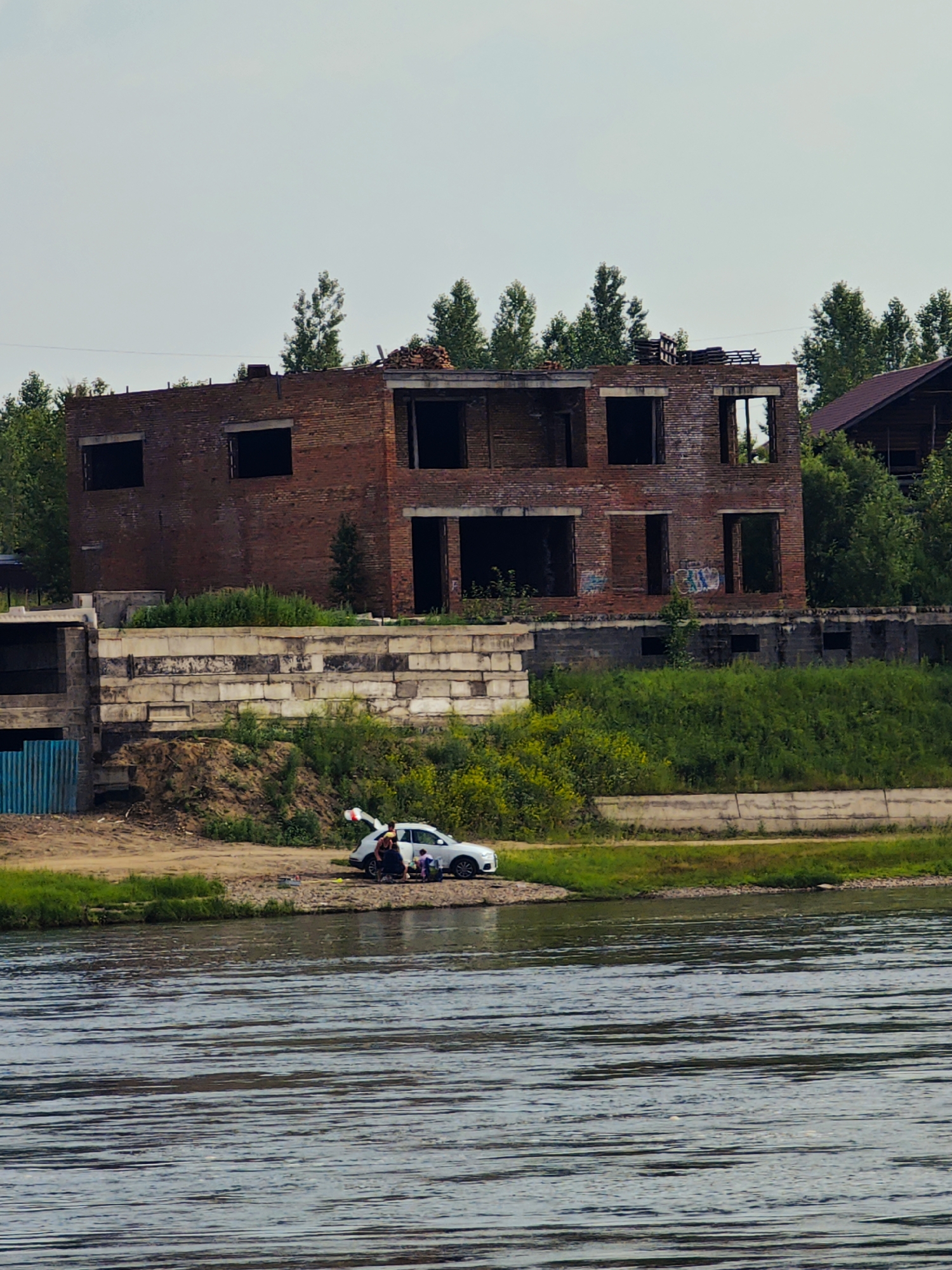 In the city of Sosnovoborsk, Russia, a partially constructed, two-story brick building stands abandoned. The building has numerous empty window and door openings, revealing a skeletal interior. Adjacent to the unfinished structure, a lower-rise building with a concrete block facade is also visible. On the dirt embankment beside a body of water, a white car is parked with its trunk open. Several individuals, appearing to be a family, are gathered near the car. One person is leaning into the trunk, possibly unpacking or packing items. Others are standing or sitting around the vehicle. Lush green vegetation covers the embankment, and trees are visible behind the structures. The sky is overcast with a light blue-grey hue.