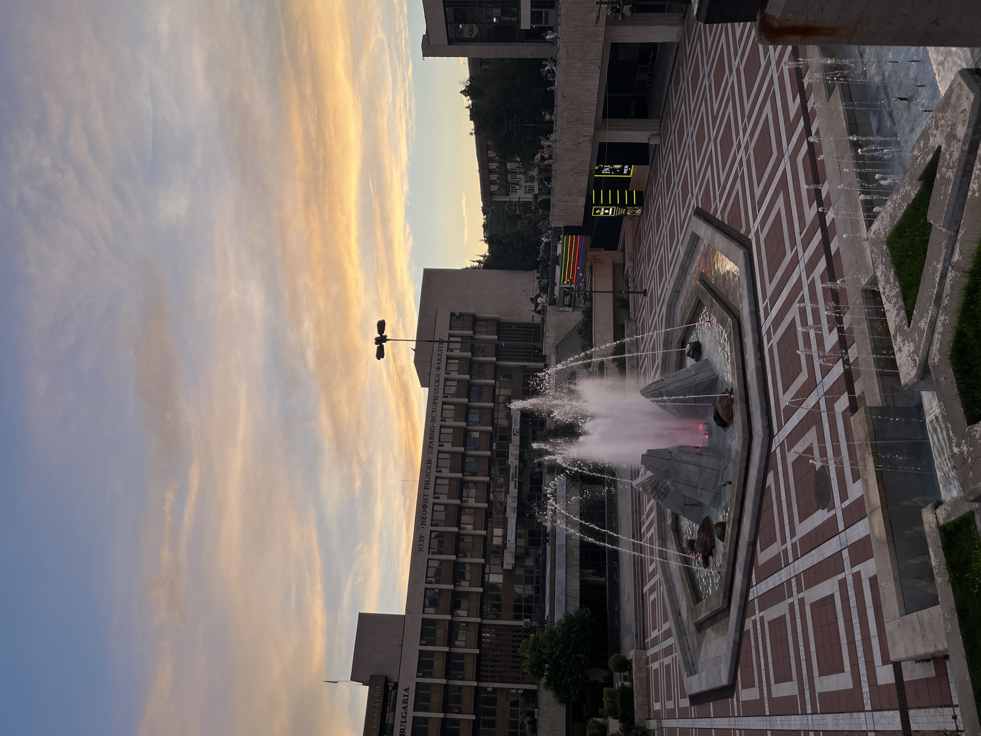 An eye-level, medium shot captures an outdoor public space with a fountain at its center under a dramatic sky filled with wispy clouds painted in hues of orange and yellow, indicative of twilight. The fountain, constructed with gray stones and illuminated with pink light, is the focal point of the foreground. It stands within a hexagonal basin. The ground is paved with interlocking geometric patterns of red and white tiles.
Dominating the background is a large, multi-story brutalist-style building with rows of windows. Text is visible on its facade, including "BULGARIA" and what appears to be Cyrillic lettering. Above this building, the sky transitions from warm oranges near the horizon to a soft blue. To the right, another building, also with multiple windows, is visible, and a balcony area with several people seated and standing is present. Pedestrians are scattered throughout the scene, some walking on the tiled plaza, others on the elevated walkway.
Further to the right, illuminated signage for "PALMS" and "DANCE" is visible. The overall environment suggests an urban setting, likely a city square or plaza, possibly near an academic institution given the visible text on the building. The lighting suggests it is late afternoon or early evening, with the setting sun casting a warm glow on the clouds. The image is taken from a slightly elevated perspective, looking down towards the fountain and the plaza.