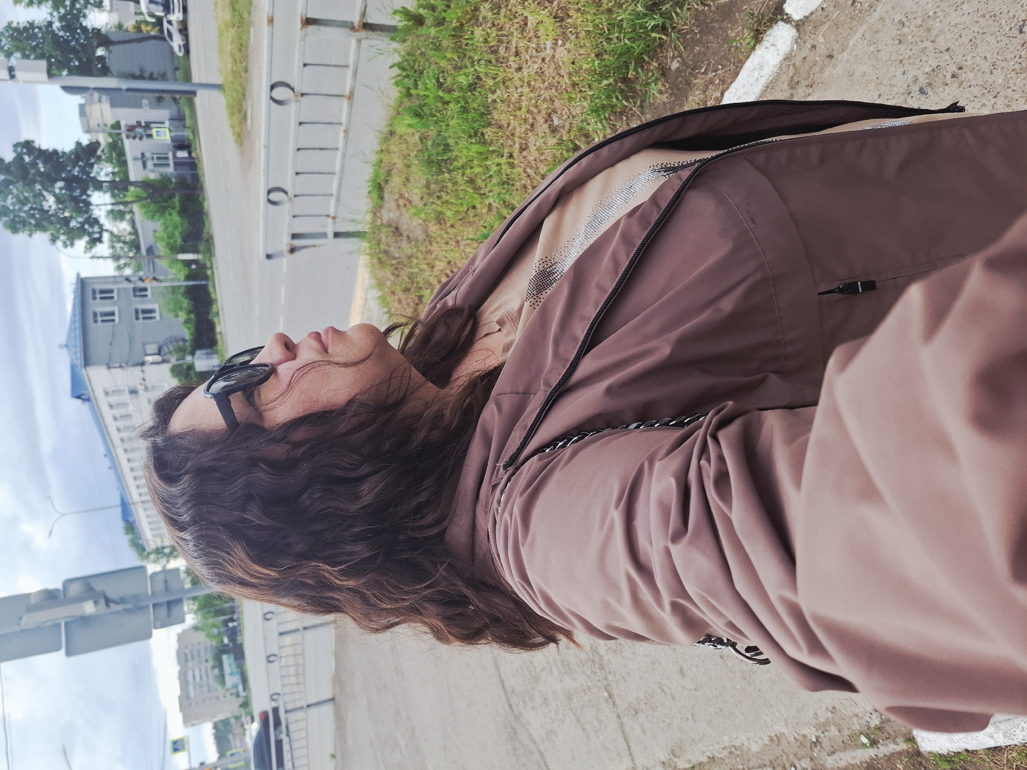 A woman with dark curly hair and sunglasses is outdoors, dressed in a brown jacket over a beige top. She is standing near a sidewalk in a streetscape. The urban setting is characterized by buildings, street signs, and pedestrian barriers visible in the background. This is in the city of Chaikovsky, Russia. The focus is on the woman and her immediate surroundings.