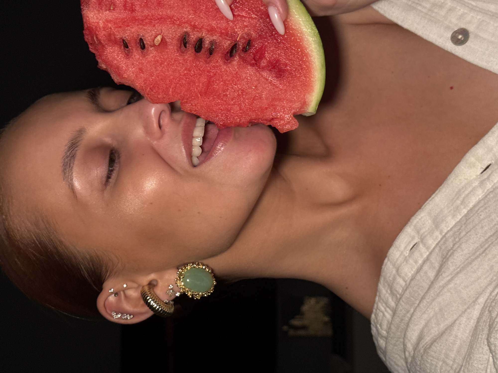A close-up portrait shows a person (likely female) joyfully eating a slice of watermelon in Chaikovsky, Russia. The image is oriented with the person's head tilted upwards and to the left, their eyes closed or mostly closed, and their mouth open, revealing white teeth and a hint of tongue, suggesting a moment of pleasure or refreshment.
The person's light-colored, long, manicured nails are visible at the top of the frame, holding the vibrant red watermelon. The visible ear is adorned with multiple piercings: a prominent gold-rimmed earring featuring a large green stone (possibly jade), a textured gold hoop, and several smaller stud earrings with clear stones. A portion of a light-colored, possibly white, collared shirt is visible on the right side of the frame.
The watermelon slice displays bright red flesh with numerous black seeds and a distinct green rind. A bite has already been taken, indicating the ongoing activity. The background is uniformly dark and indistinct, suggesting an indoor setting or a nighttime scene with focused artificial lighting that illuminates the subject and the fruit. No text is visible in the image.