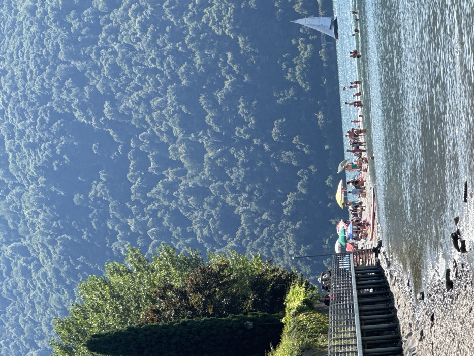 The image presents a sunlit, crowded pebble beach scene on Lake Como, specifically in Abbadia Lariana, Italy. In the foreground, calm, reflective lake water transitions to a narrow, rocky shoreline. Extending from the left is a raised wooden pier or walkway with metal railings, supported by vertical posts, and bordered by dense green foliage, including a tall, dark conical tree. A streetlamp is visible next to the walkway.
The midground features a busy beach area densely populated with individuals of varying ages, including adults and children. Numerous people are standing, sitting, or lying on the sand, with several colorful beach umbrellas providing shade (including yellow, blue, and red umbrellas). A red and white paddleboard or small boat rests on the beach near the water's edge. Many individuals are observed wading or swimming in the shallow, clear lake water adjacent to the beach. Further out on the lake, to the right of the beach, a white-sailed sailboat moves across the water.
The background is dominated by a large, steep mountain face densely covered in green trees and foliage, appearing misty in the distance under a clear, bright sky.