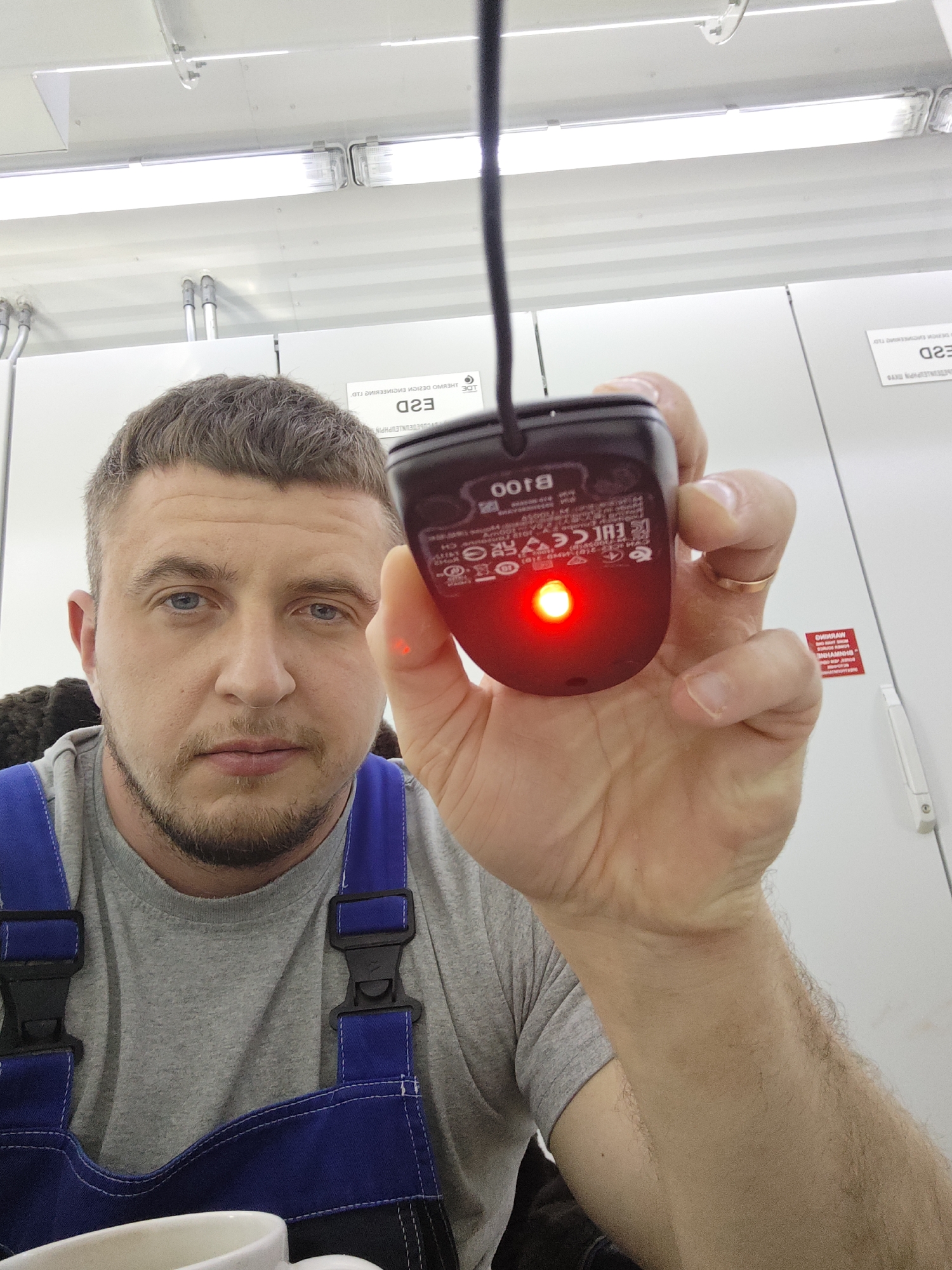 A man with a beard is holding up a black B100 computer mouse with a red light emanating from the bottom in a bright, nondescript interior location. He is wearing a grey t-shirt and blue overalls. White walls and ceiling with fluorescent lighting are visible in the background, as well as some labels/signs with text and symbols. A coffee mug is slightly visible at the bottom. The location is Unknown, Unknown.