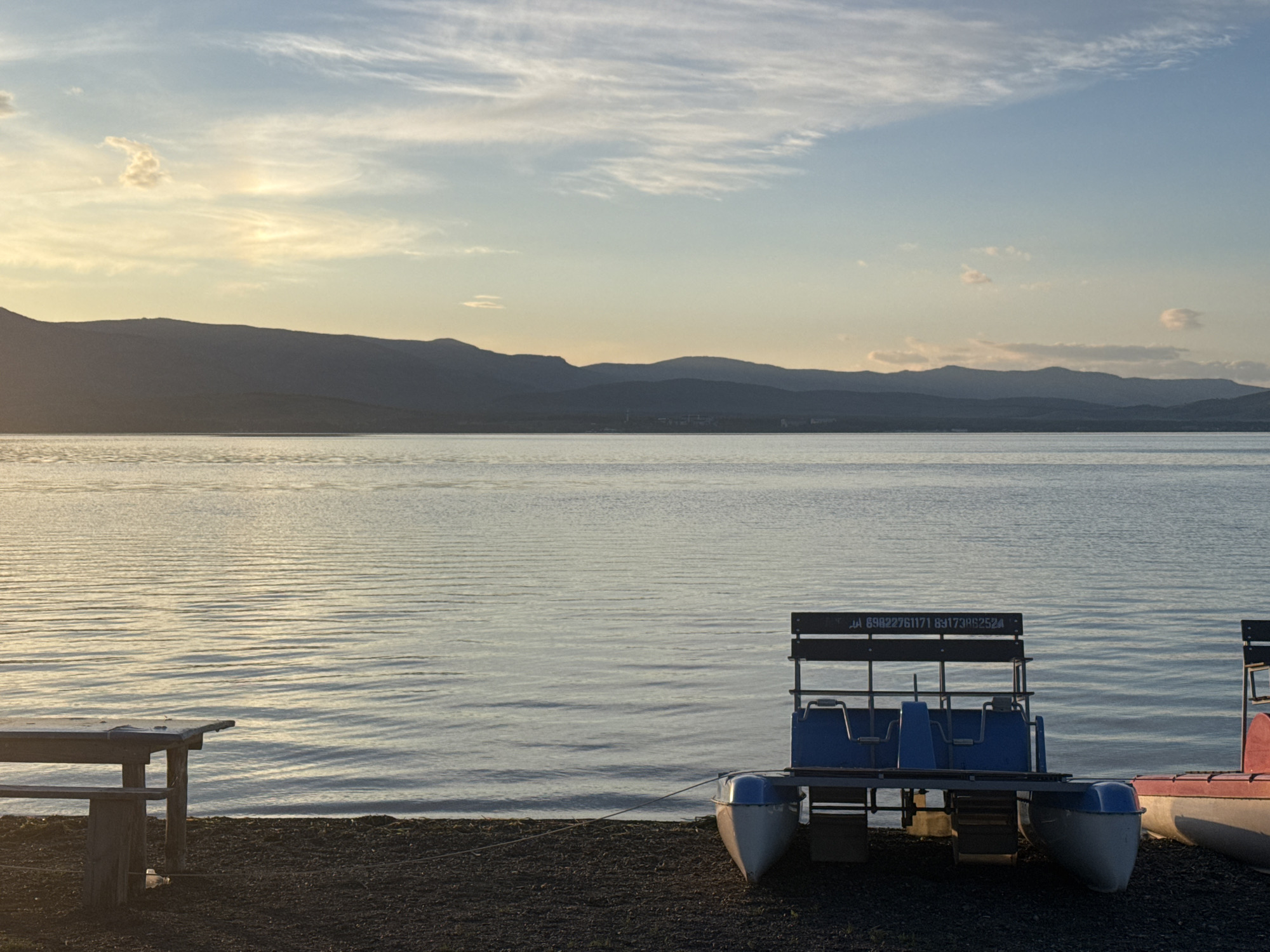 The image depicts a tranquil lake scene at dusk in the city of Yakty-Kul, Russia. In the foreground, a dark-colored ground with small pebbles leads to the calm water. On the left side, a rustic wooden picnic table with a bench is positioned near the shore. To the right, a blue and white pedal boat is tethered to the shore, with visible text on its backrest reading "JA 69822761171 69173852524". Another partially visible red and white boat is to the far right. The lake surface reflects the soft light of the setting sun, with gentle ripples creating a shimmering effect. In the background, a range of silhouetted mountains stretches across the horizon, softened by a hazy atmosphere. The sky above is a blend of pale blue and warm yellow hues, with wispy white clouds scattered across it, suggesting a serene end to the day. There are no people visible in the image.