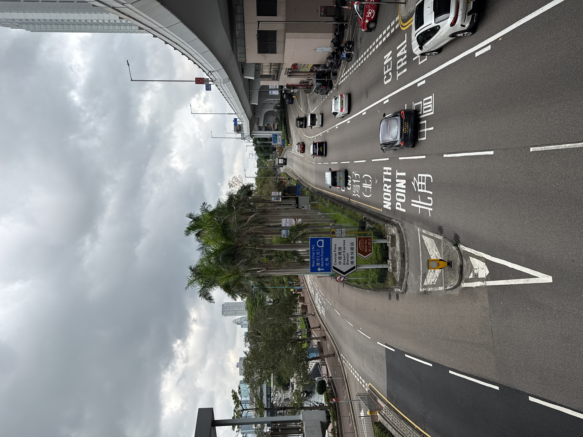 The image captures a view of a multi-lane roadway in Hong Kong, China. The roadway features designated lanes with painted markings indicating "NORTH POINT (北角)" and "CENTRAL (中環)". Multiple vehicles are present, including cars and taxis. Overhead is a highway overpass. Palm trees line the side of the road, and signs indicate directions to Wan Chai (N), North Point, Central Piers, and the Airport Express Station. The sky is overcast with grey clouds. The perspective suggests the image was taken from an elevated position.