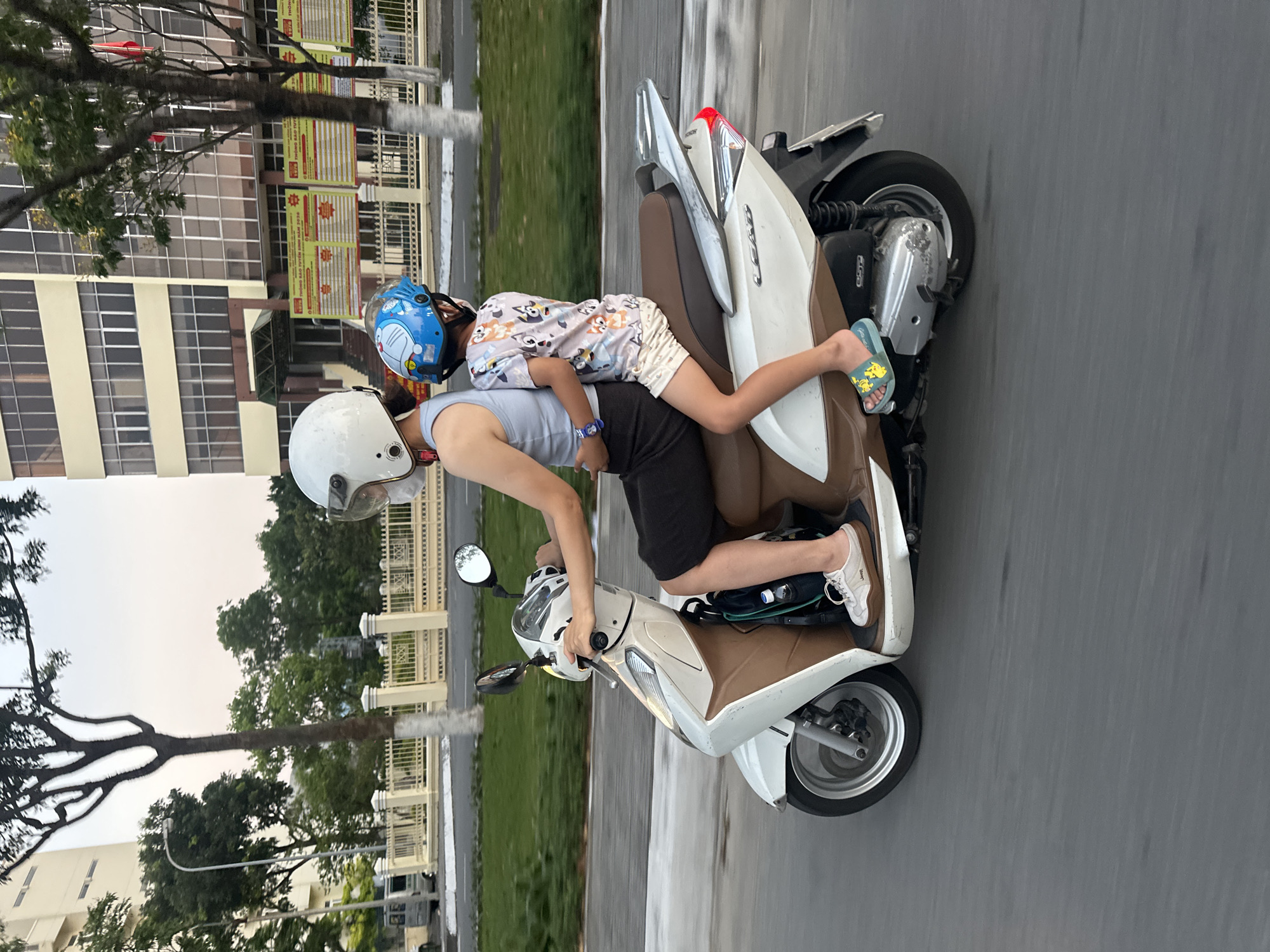 A person wearing a white helmet rides a white and brown scooter down a gray asphalt road, with a child lying prone on the seat behind them, also wearing a blue helmet. The child is wearing patterned pajamas and sandals. The adult rider wears a light-colored top and dark shorts, with white sneakers. Trees and buildings are visible on either side of the road. The location is Đà Nẵng, Vietnam.