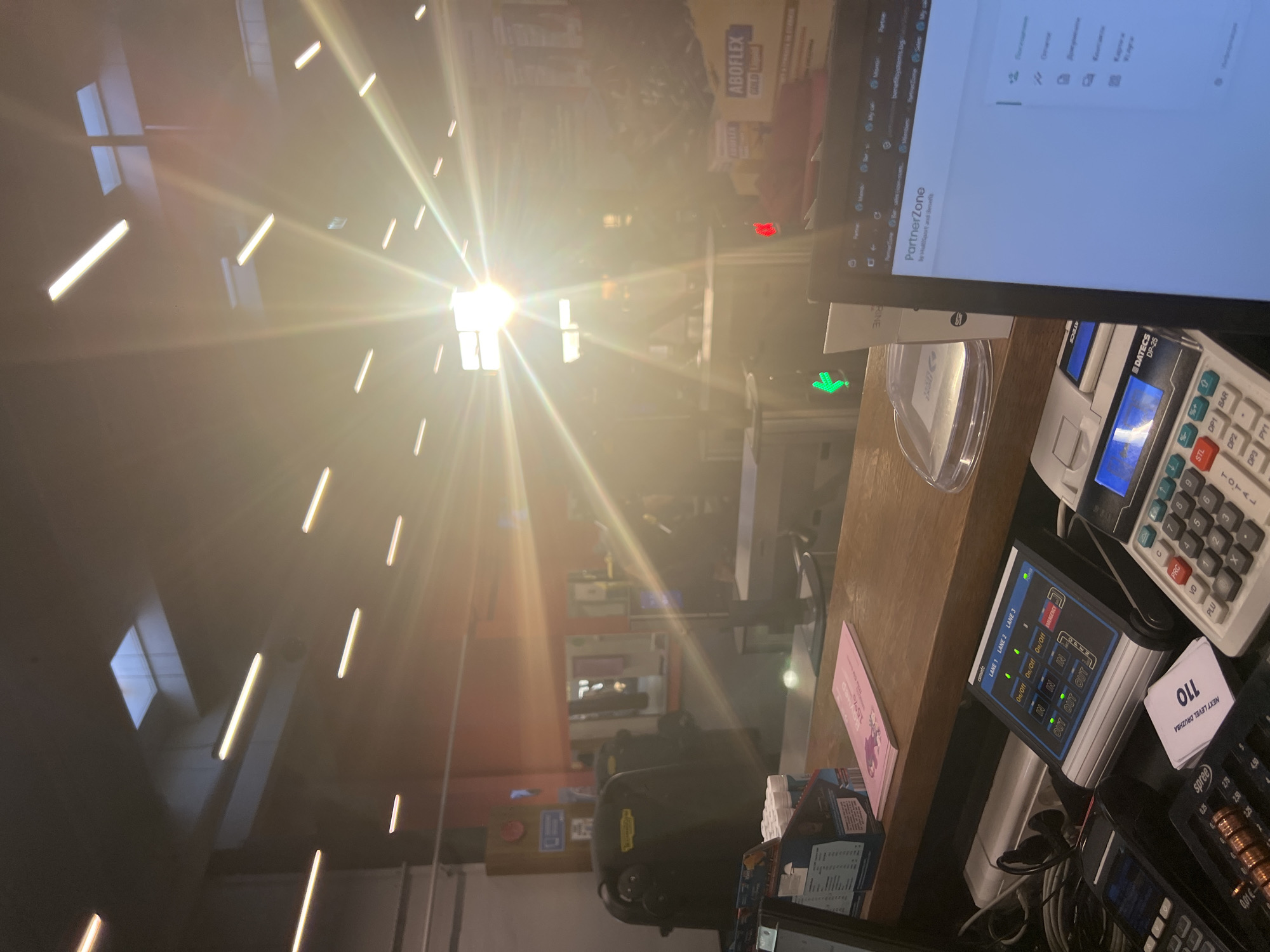 Here's a detailed description of the image:
**Location:** The photo appears to be taken from the perspective of a cashier at a store in Sofia, Bulgaria, likely a construction or hardware store based on the products visible on the computer screen and in the background.
**Environment:** The interior has bright overhead lighting, possibly with sunlight filtering through windows or skylights, creating a strong glare in the center of the image. The surrounding area consists of shelves stocked with merchandise.
**Objects:**
* **Point of Sale (POS) System:** A digital monitor displays software interface labeled as "PartnerZone" by "MultiSput and Benefit". There are various POS peripherals like a calculator (Datecs DP-25), payment terminal with an active screen, and a lane management system.
* **Cash Register Area:** The counter has a light wooden surface and contains various items including receipts, a plastic coin cup, and promotional materials, with items like candies or small goods displayed.
* **Signage:** A small card with the number "110" is visible, likely a ticket number for a queuing system, along with the words "NEXT LEVEL DRUZHBA".
**Actions & Interactions:**
* The scene suggests a point-of-sale transaction in progress, with the cashier side of the counter clearly visible. The state of the screens implies that the system is active and ready for input.
**Subjects:**
* While there aren't any people prominently visible, the presence of the cash register and POS system suggests that a cashier and customers are part of the broader scene.
**Additional Notes:**
* The brand "ABOFLEX GOLD" is visible on a product in the background, potentially an item sold in the store.
* The payment terminal displays text like "LANE 1 LANE 2 LANE 3", and button functions like "On/Off", "In/Out" and "Emergency" suggesting some type of monitoring system is in place.