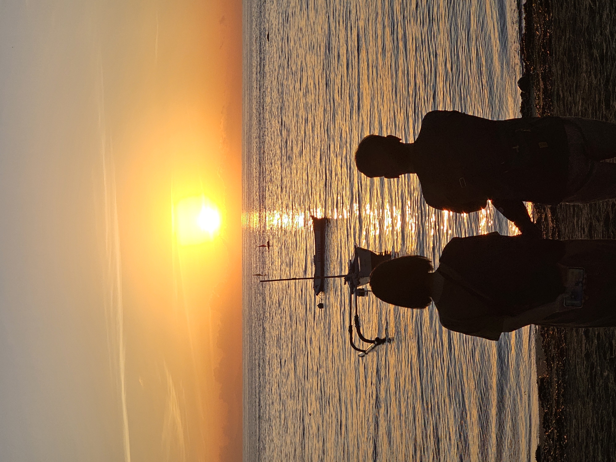 Two people stand silhouetted against a sunset backdrop on a beach in Denpasar, Indonesia. The sun is a bright orange orb on the left, casting a golden hue across the water and sky. A small boat is visible in the water, its details obscured by the light. The silhouettes of the individuals stand to the right, adding depth to the scene. The beach is visible as a dark strip on the right edge of the frame.