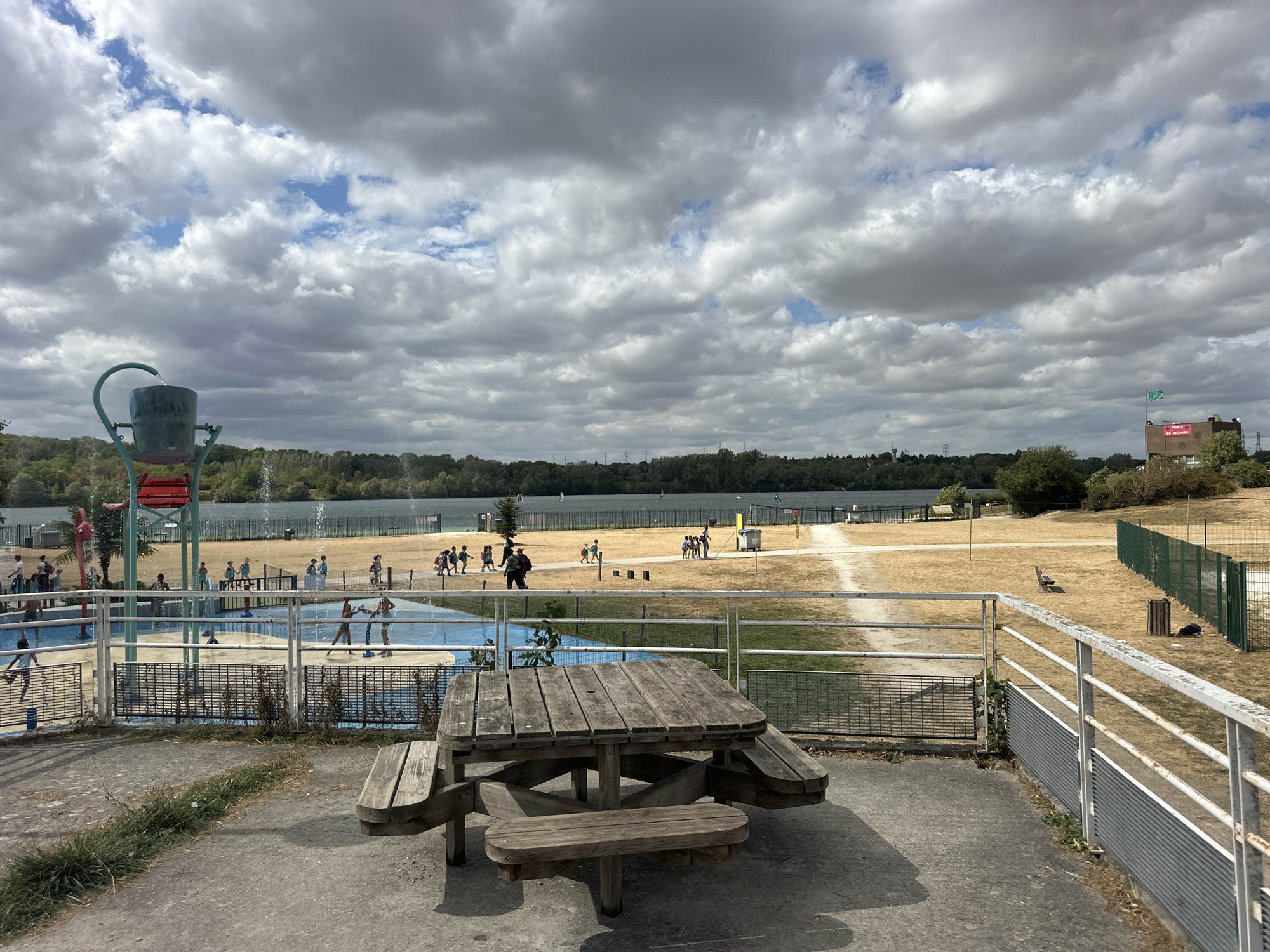 The image depicts an outdoor scene at the Jablines-Annet lake in Jablines, France, on a cloudy day. In the foreground, a weathered wooden picnic table with attached benches sits on a concrete patio. A metal railing runs along the edge of the patio, overlooking a water play area for children. This area features blue and yellow surfaces with water fountains and several children playing. Beyond the water area is a sandy beach, and further back, a large body of water (the lake) is visible, with some windsurfers. The background consists of a treeline and buildings, including one with a red banner and a green flag. The overall environment appears to be a recreational park or leisure area.