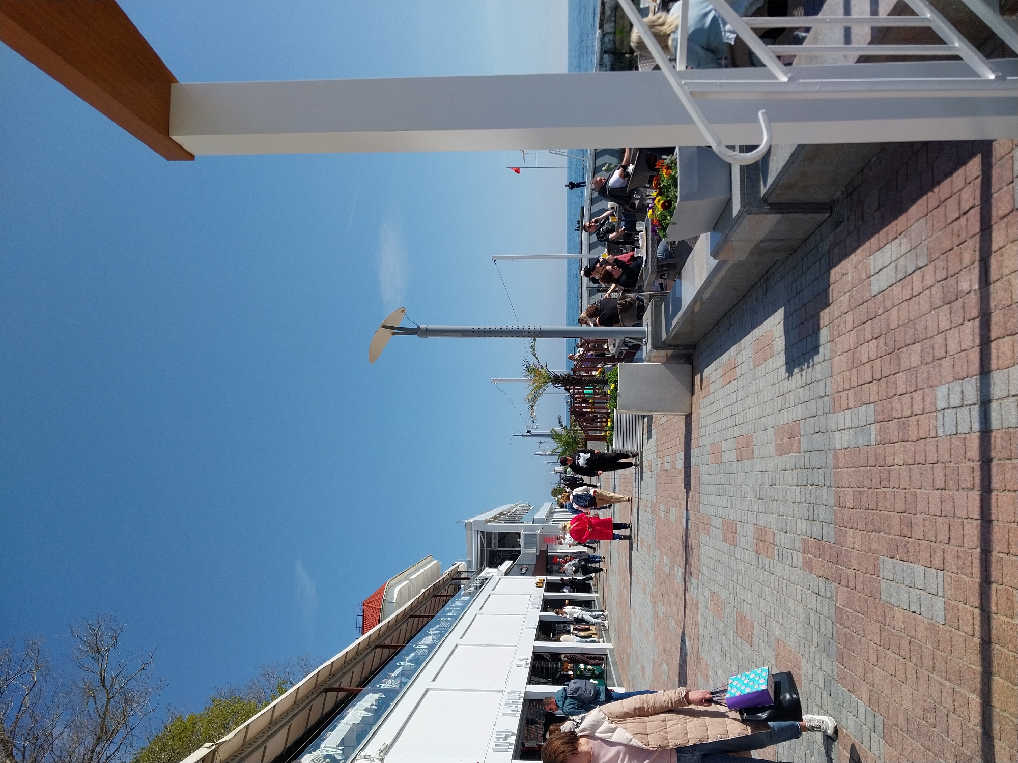 This image captures a lively outdoor promenade or boardwalk alongside a large body of water under a bright, clear blue sky. The scene is bathed in strong daylight, casting distinct shadows, indicating a sunny day.
In the foreground, a prominent white column supports a thick, light brown wooden beam, likely part of an overhang or architectural structure. The ground is paved with a distinctive pattern of alternating reddish-brown and grey rectangular blocks. A person is partially visible in the bottom right, holding a colorful blue and purple patterned shopping bag and a black handbag, walking towards the right.
Along the promenade, numerous people are engaged in various activities. Many are seated at outdoor tables under white canopies or umbrellas, suggesting cafes or restaurants, enjoying the waterfront view. Others are strolling along the paved walkway. A tall, modern lamppost with a curved, wing-like shade stands in the mid-ground.
To the left of the promenade, a long, white modern building with large glass windows stretches along the path, housing what appear to be shops or dining establishments. Further down, another building with a reddish-orange tiled roof is visible. Green trees and foliage are present further back. In the distance, beyond the people and buildings, the calm blue water meets the horizon. A small red flag is visible on a pole near the water's edge in the background.
Visible text on the building facades in the mid-ground is largely unreadable due to distance and angle, but some stylized patterns are visible on glass barriers. The overall atmosphere appears leisurely and pleasant.
