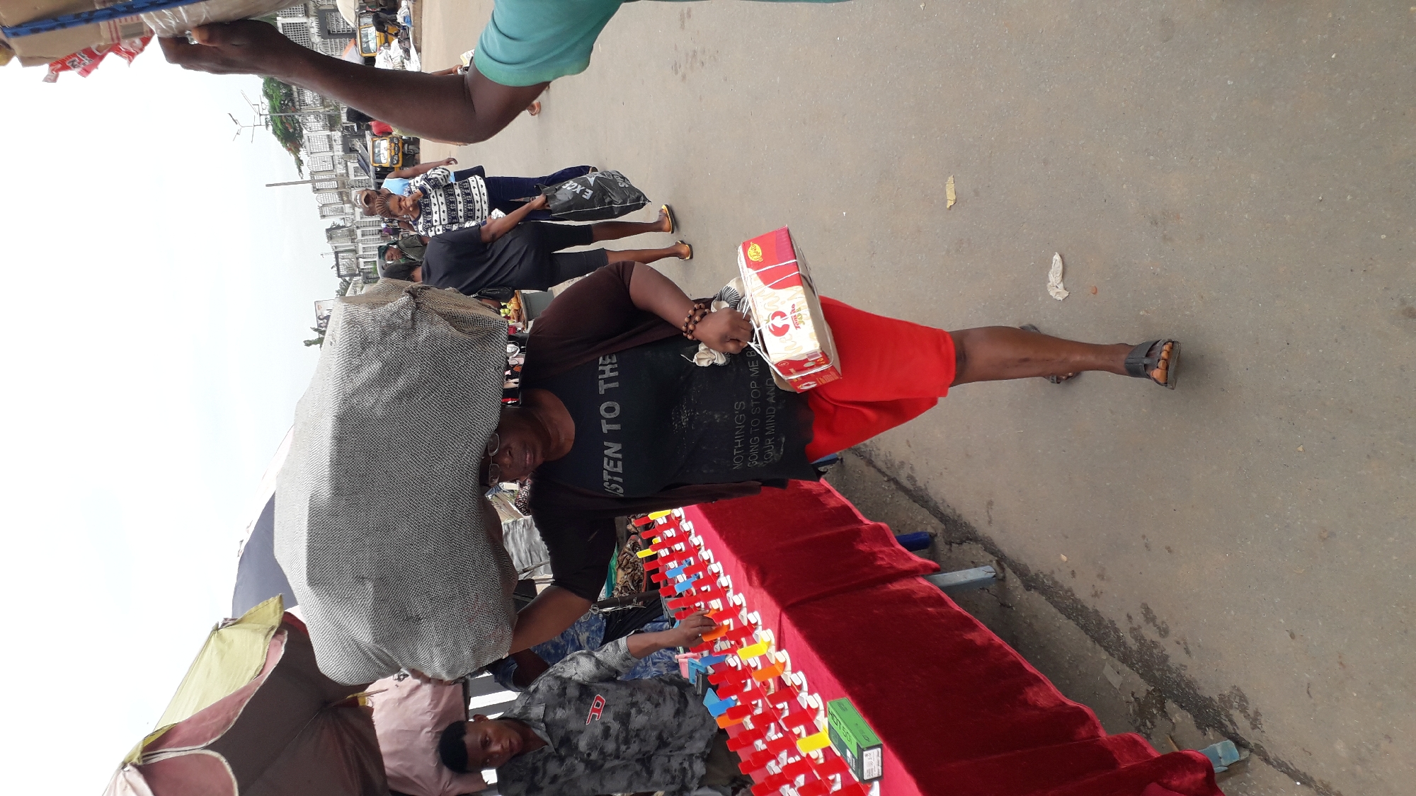 The image captures a street scene in Onitsha, Nigeria. A woman wearing red shorts, a dark t-shirt, and sandals stands beside a table covered with a red cloth, on which items for sale are arranged. She holds a cardboard box in her hand. A large bundle is on her head. A man in a grey patterned shirt rests on the ground near the table. Other people can be seen walking in the background on the road. Umbrellas and building facades are visible in the background. The environment appears to be a bustling market area. The road surface is paved.